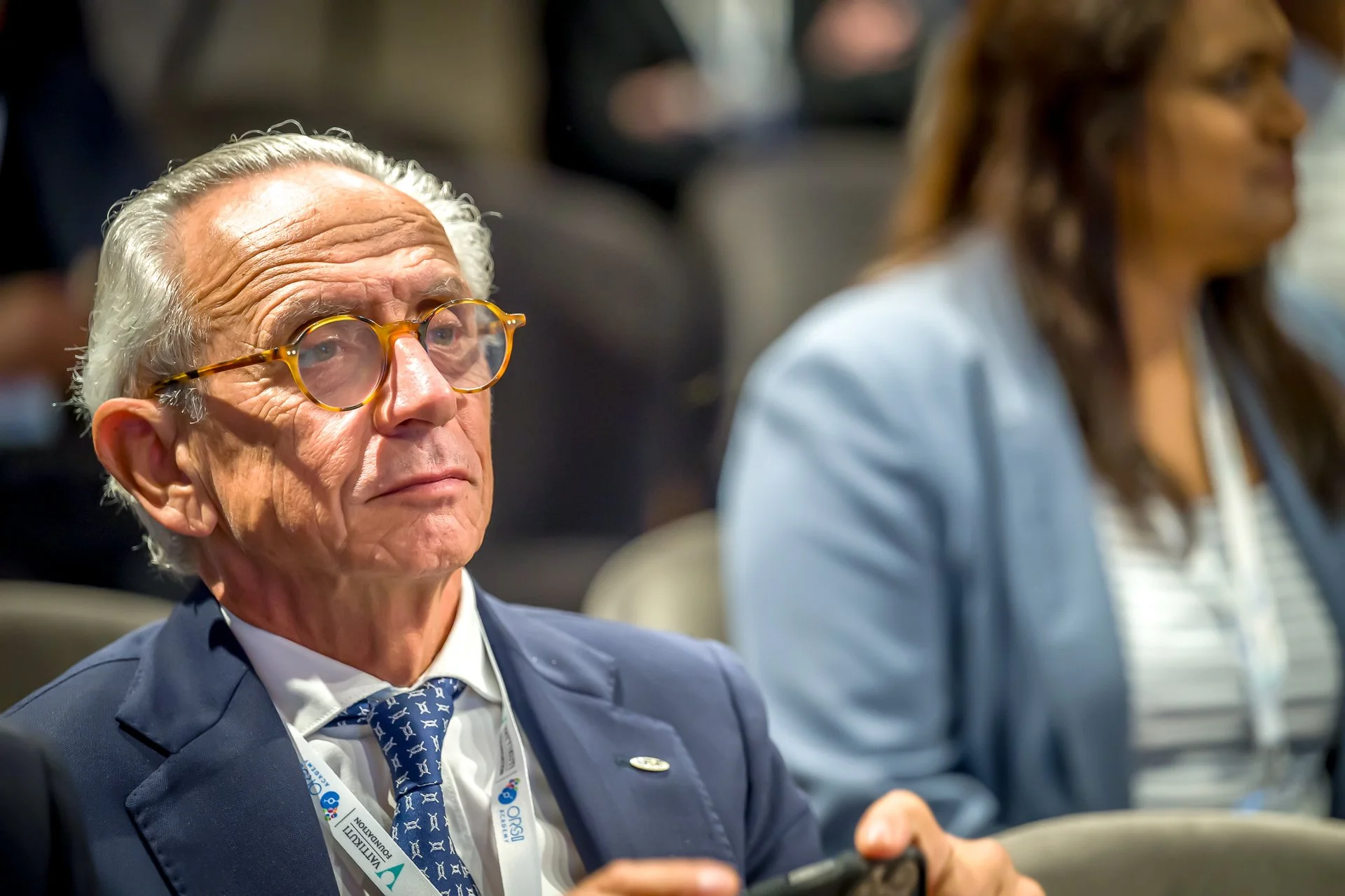 Un homme âgé avec des lunettes assis dans une salle de conférence, attentif, portant un costume bleu et une cravate.