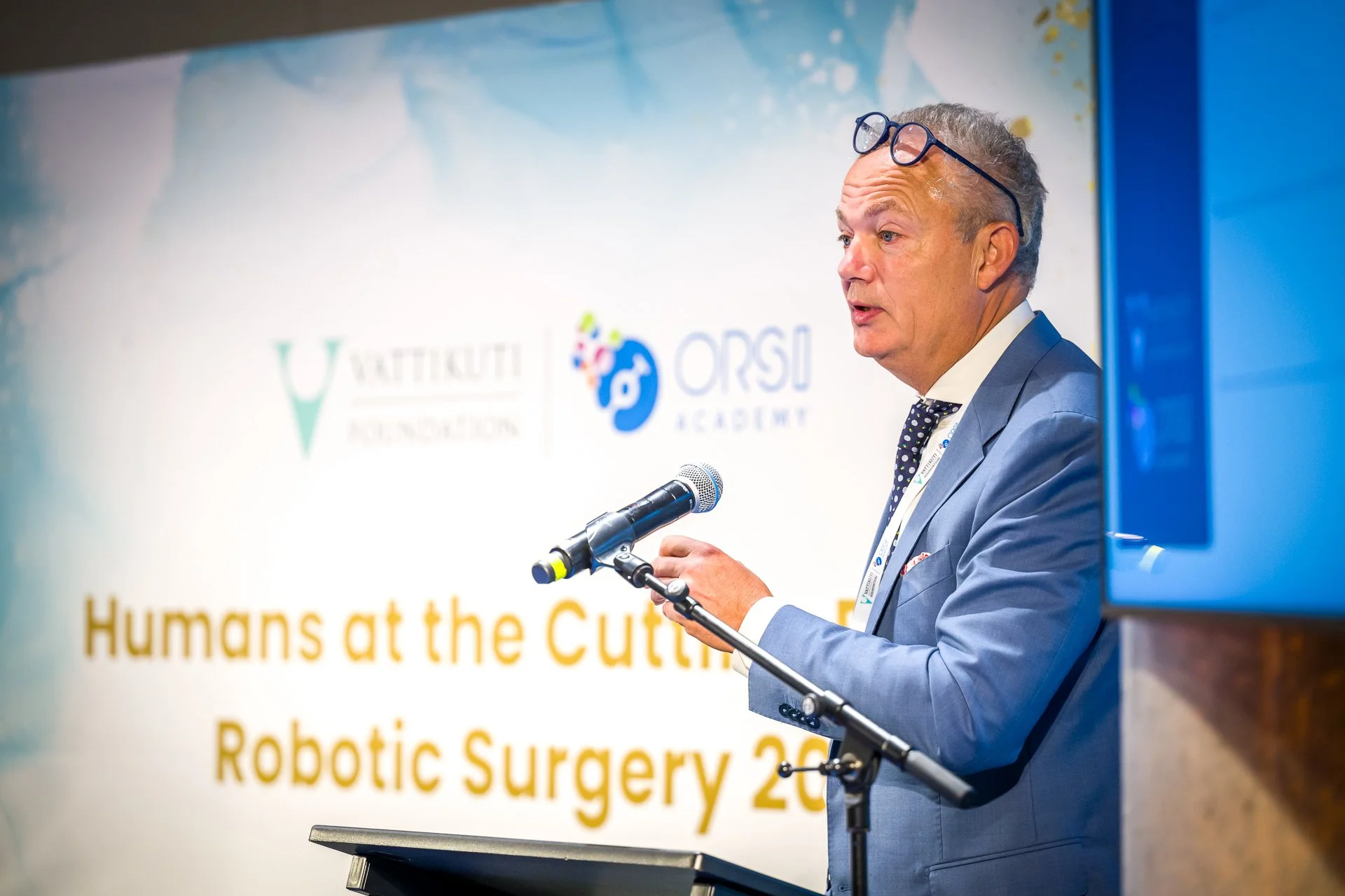 Un homme en costume bleu parle devant un micro lors d'une conférence sur la chirurgie robotique, avec des logos de la Vattikuti Foundation et ORSI Academy en arrière-plan.