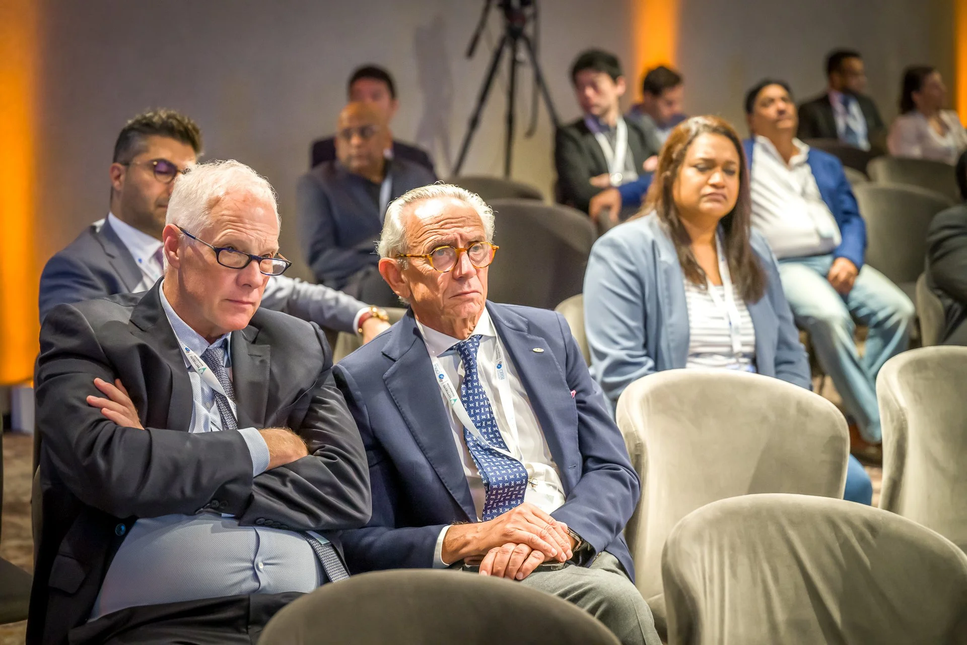 Personnes assises lors d'une conférence, portant des costumes, dans une salle avec d'autres spectateurs.