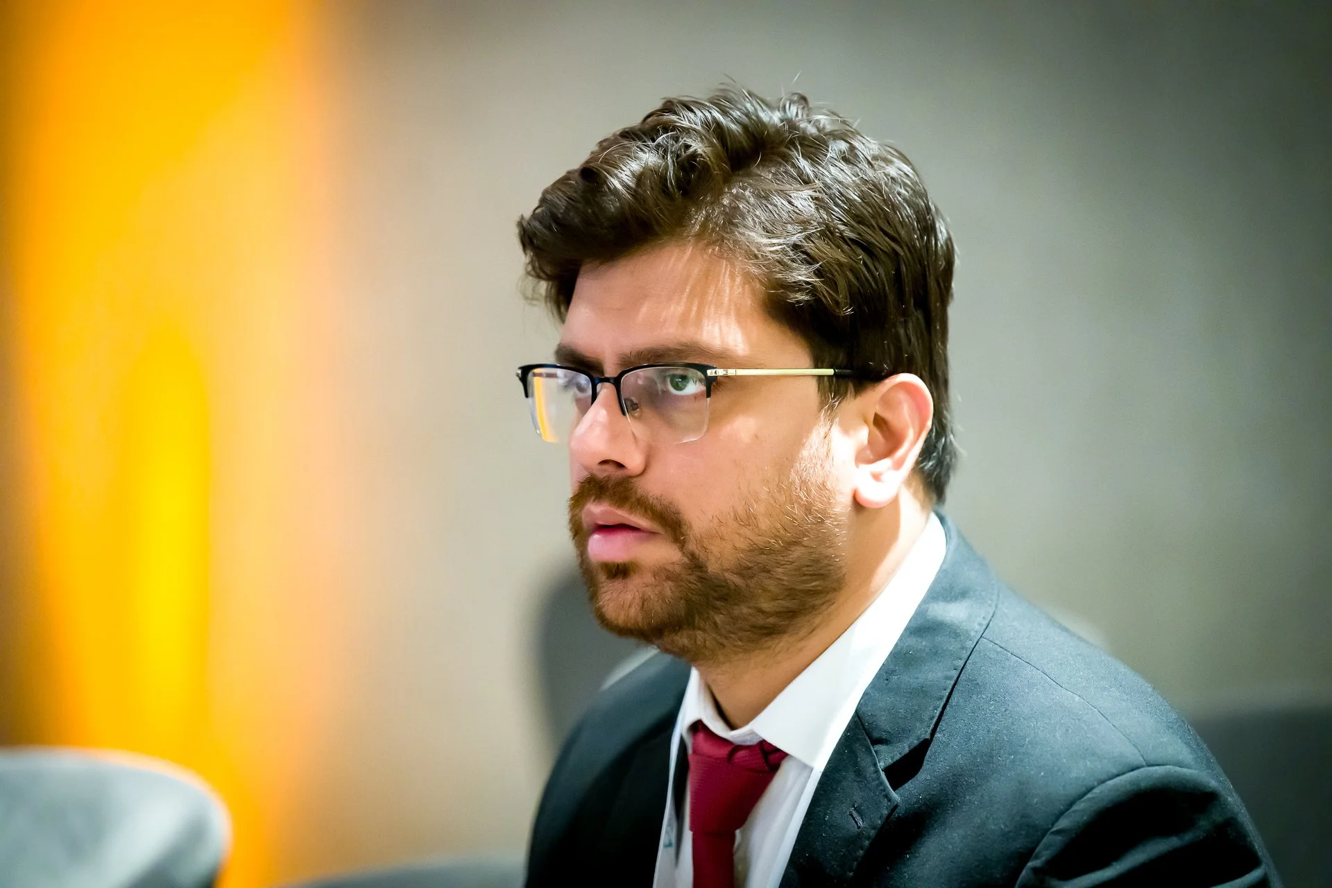 Un homme portant des lunettes et un costume noir avec une cravate rouge, regardant vers la droite de l'image.