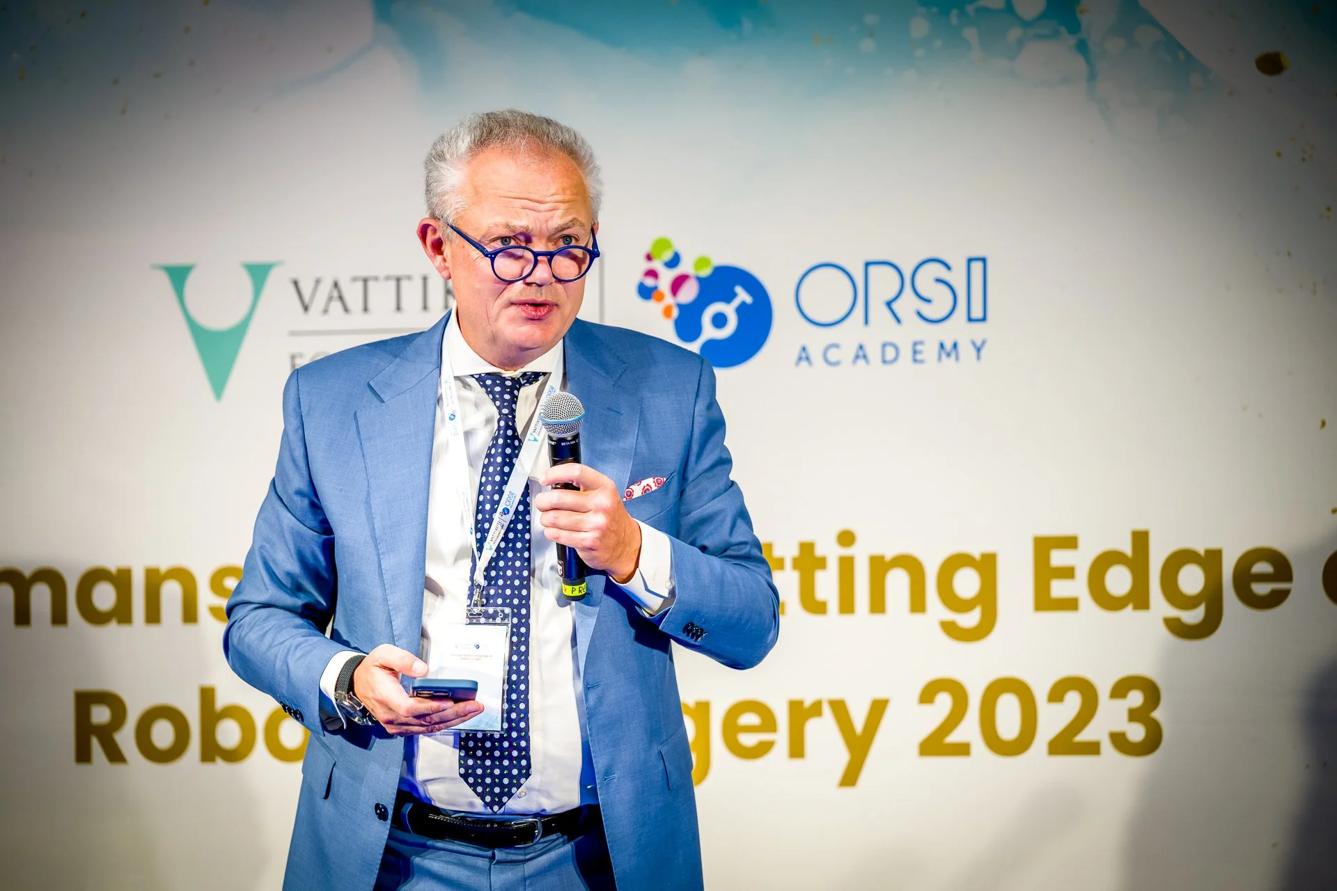 Homme en costume bleu avec un microphone sur une scène de conférence, logos "VATTIKUTI" et "ORSI Academy" en arrière-plan.