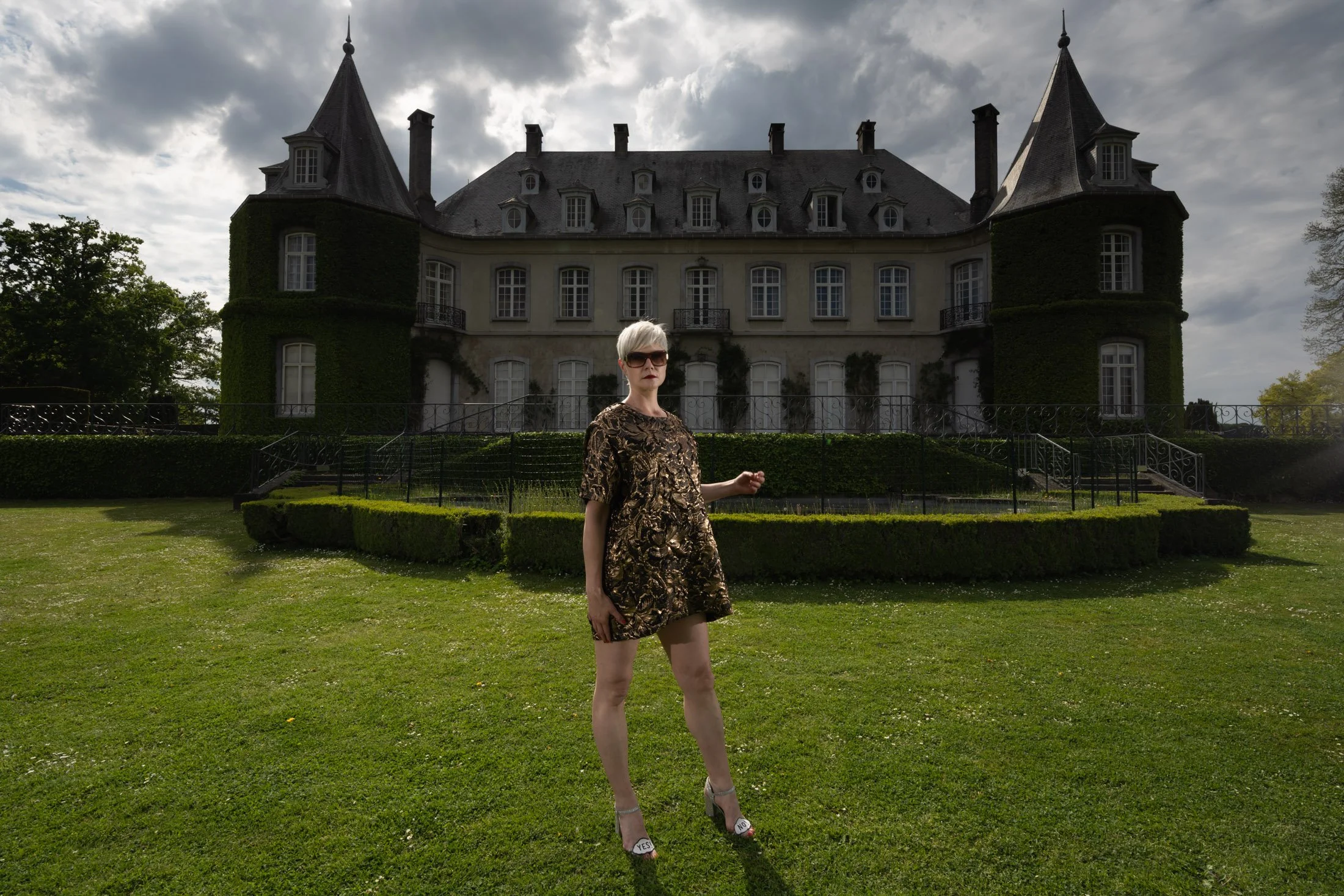 Une personne posant devant un grand château avec des tours et une façade recouverte de lierre, dans un jardin verdoyant.