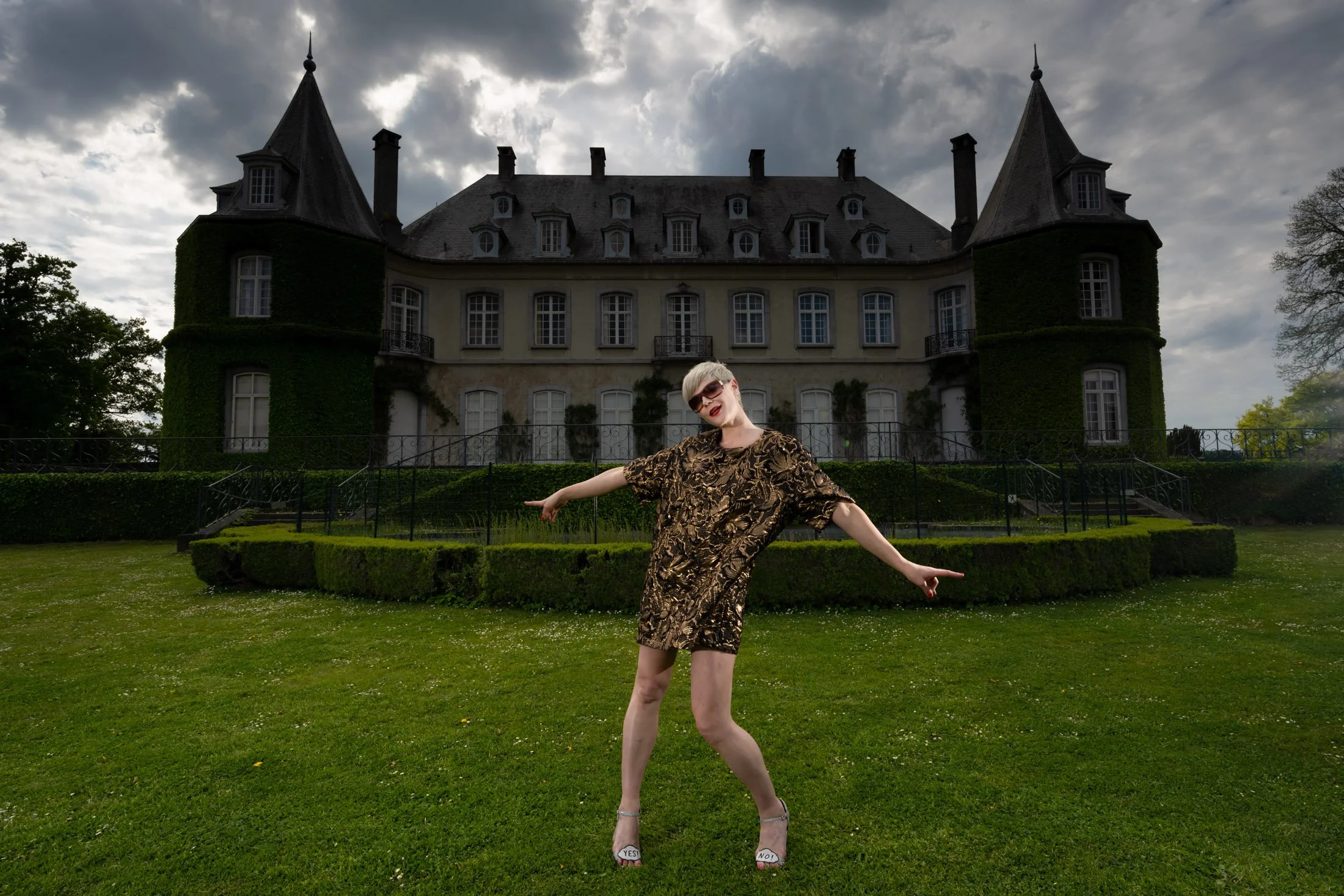 Personne souriante posant devant un grand manoir avec des tourelles, espace vert autour.