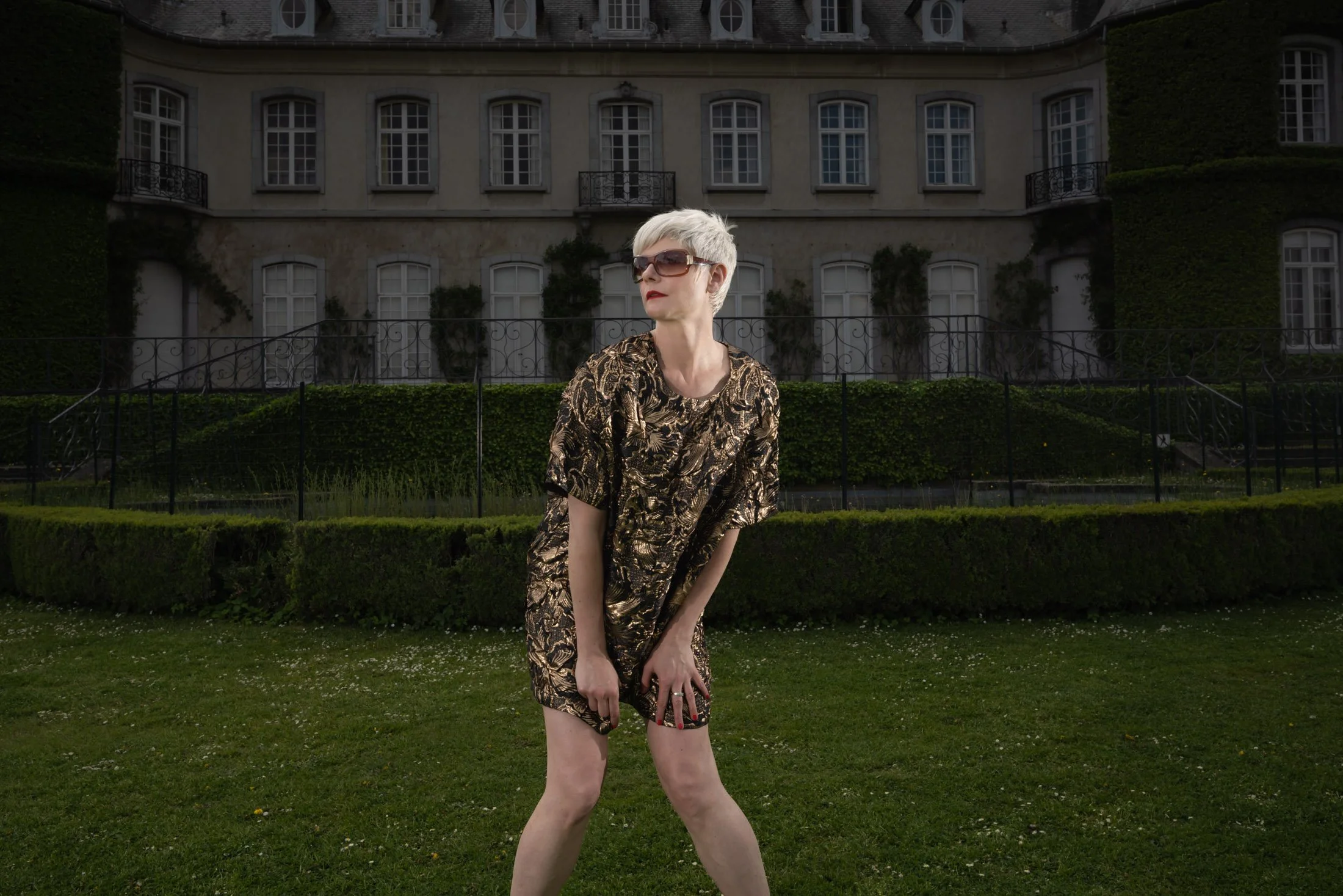 Une personne aux cheveux courts et blancs, portant une robe à motifs, pose dans un jardin devant un grand bâtiment ancien avec des fenêtres françaises et du lierre.