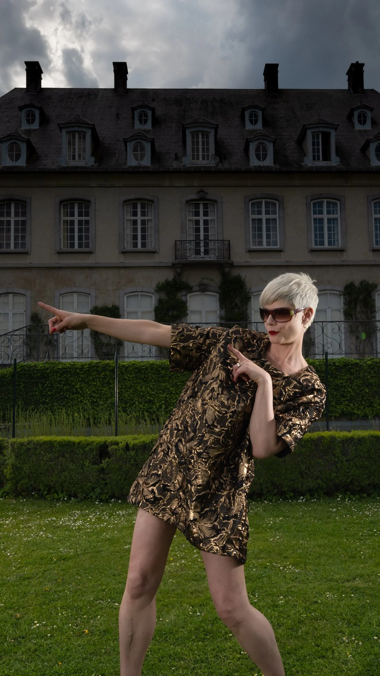 Femme en robe dorée devant une grande maison, posant avec un geste dynamique.