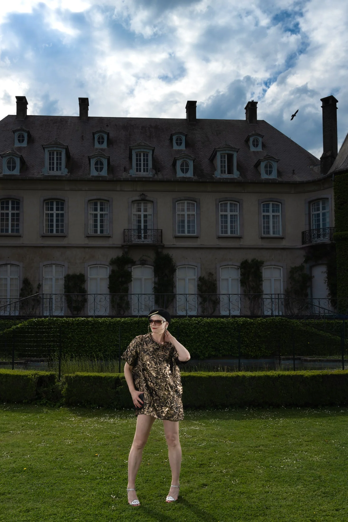 Une personne posant devant un grand bâtiment ancien, avec un ciel nuageux.