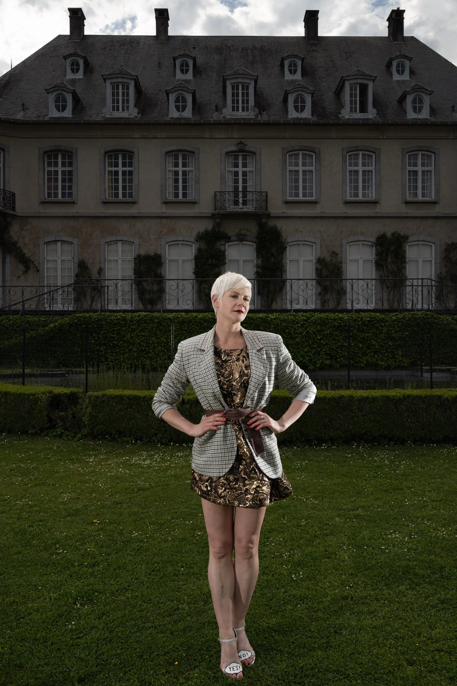 Une personne pose devant un grand bâtiment ancien avec de nombreuses fenêtres. Elle porte une tenue élégante avec une veste à carreaux et une robe dorée, et ses chaussures ont la mention "Yes" et "No." Le bâtiment est entouré de verdure.