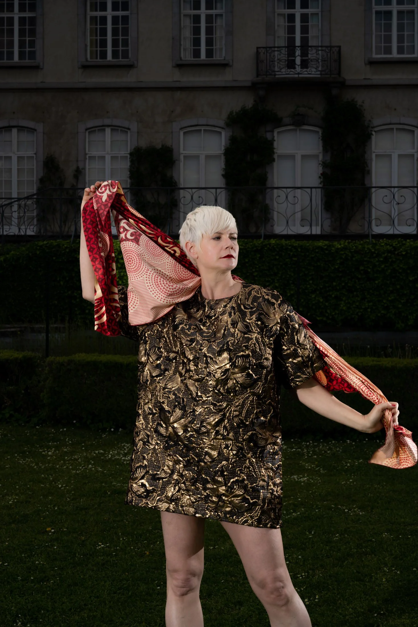 Une personne aux cheveux courts blancs, portant une robe à motifs dorés, tient un foulard coloré devant un bâtiment ancien et un jardin.