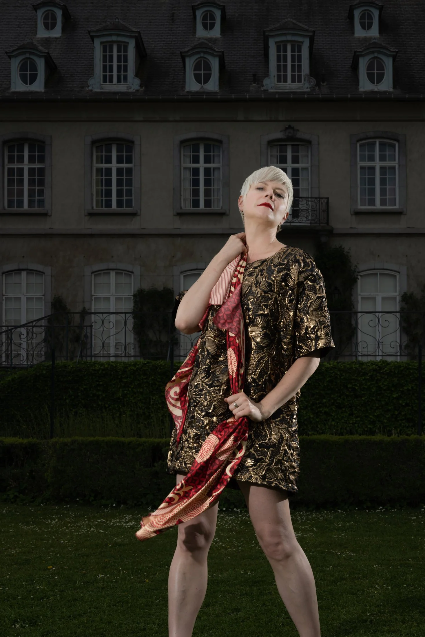 Une personne avec des cheveux courts blonds pose élégamment devant un grand bâtiment classique avec des fenêtres mansardées. Elle porte une robe à motifs dorés et tient un foulard rouge et doré.