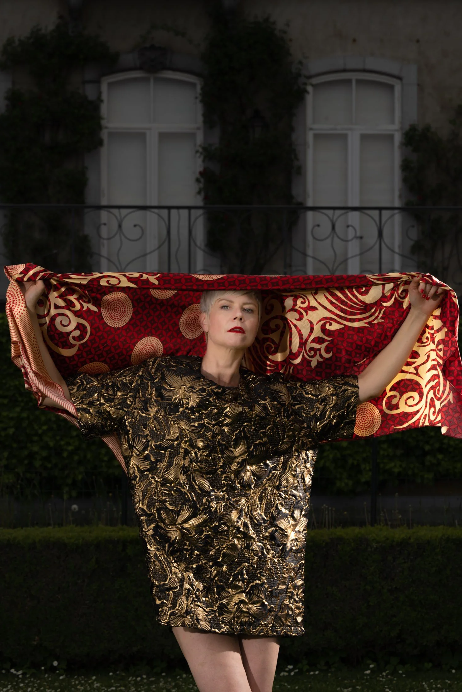 Femme debout dans un jardin, vêtue d'une robe dorée et tenant un tissu rouge avec des motifs dorés et noirs au-dessus de sa tête. Derrière, il y a une maison avec des fenêtres et des plantes grimpantes.