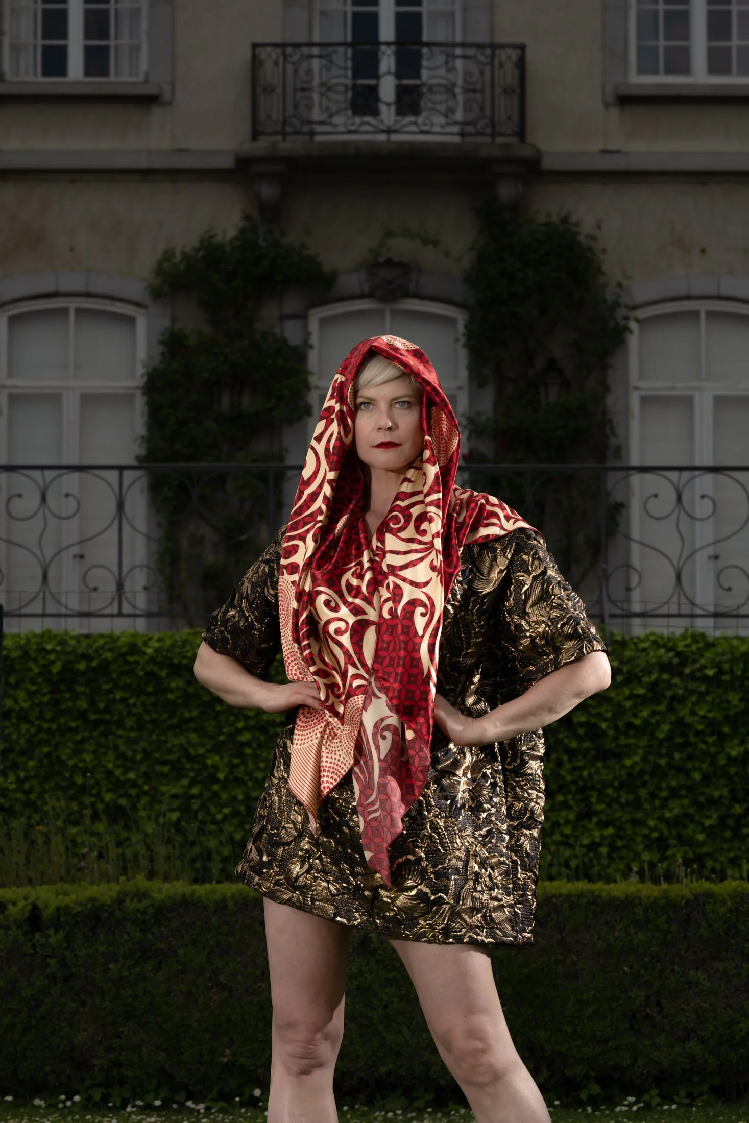 Femme posant avec un foulard rouge et or devant un bâtiment ancien.