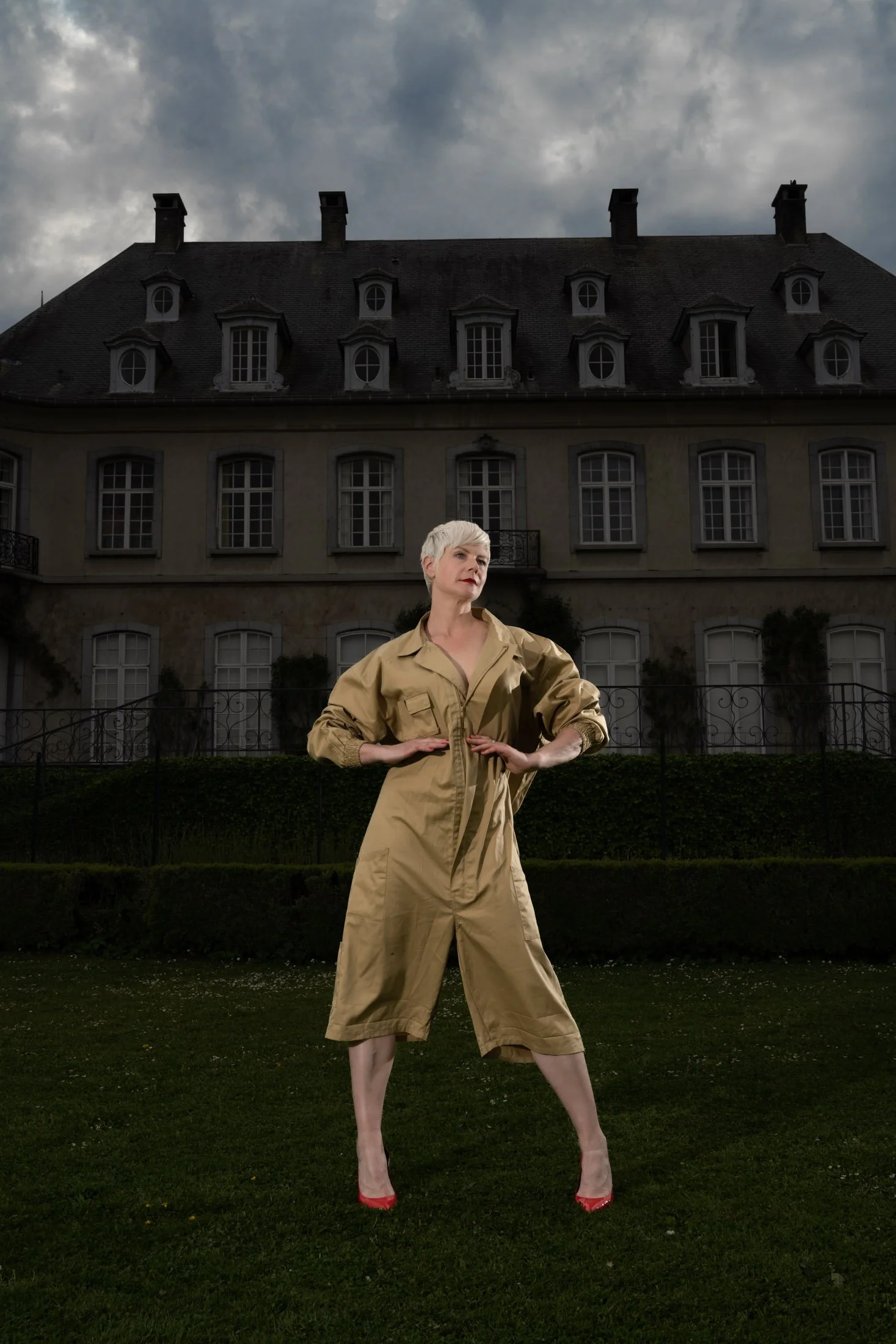 Personne en combinaison beige devant un manoir, ciel nuageux en arrière-plan.