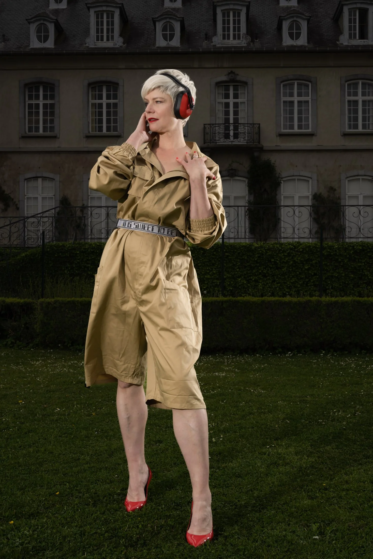 Personne portant des écouteurs rouges, manteau beige, talons rouges, devant un bâtiment ancien.