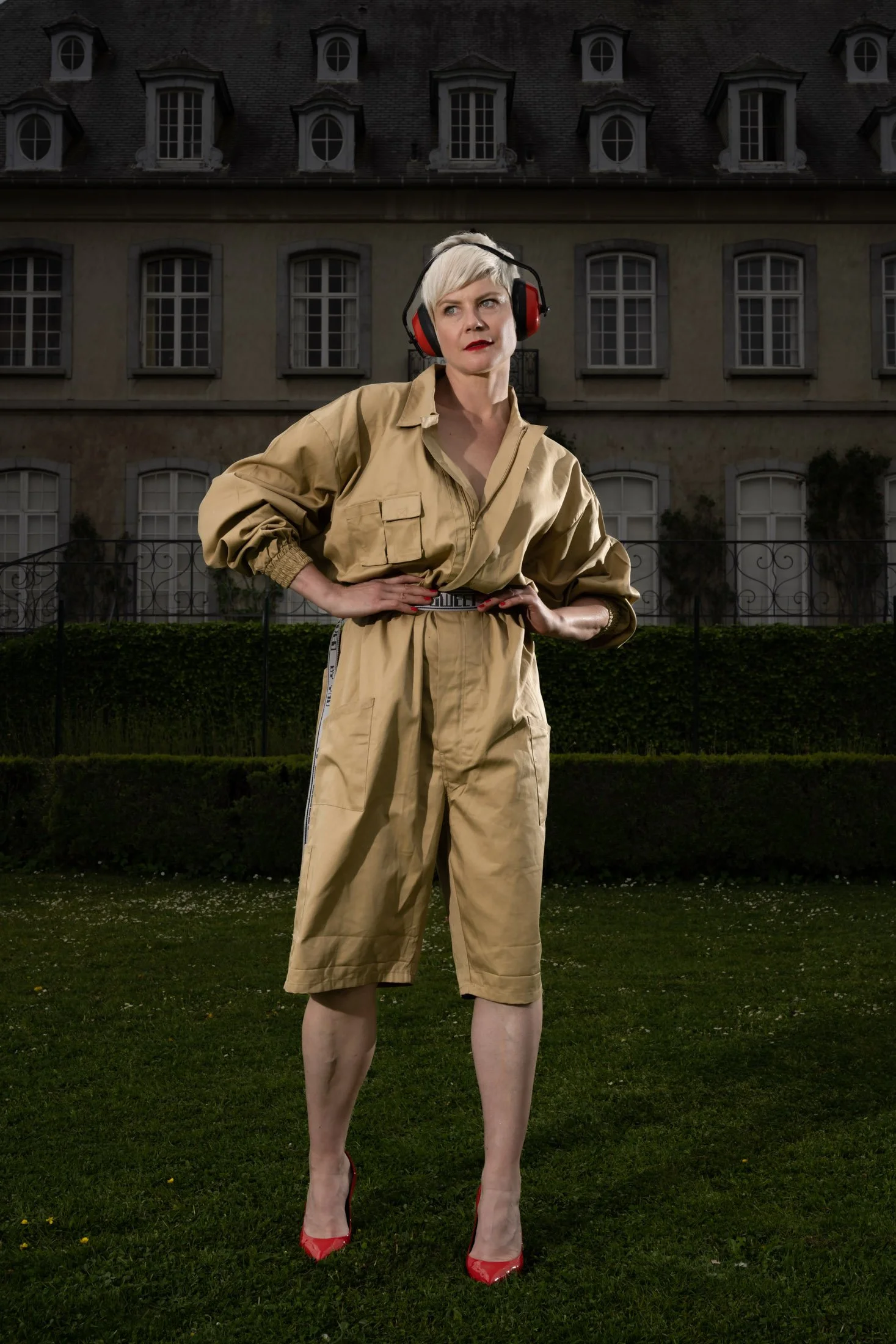 Personne portant une combinaison beige, des protections auditives rouges et des talons rouges, posant devant un bâtiment classique avec des fenêtres en arrière-plan.