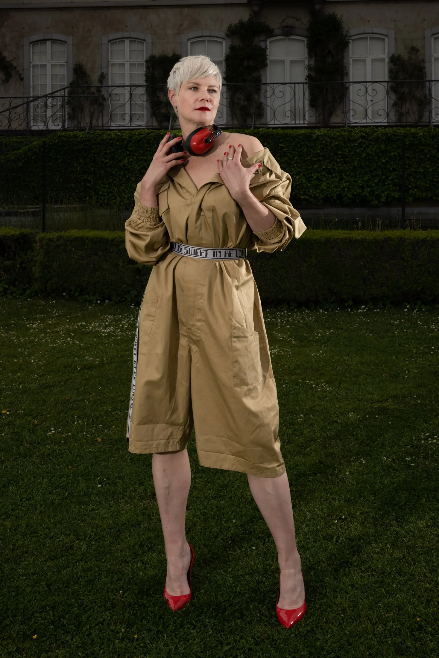 Femme avec coupe courte blonde, portant un manteau beige et des talons rouges, se tenant sur une pelouse devant un bâtiment.