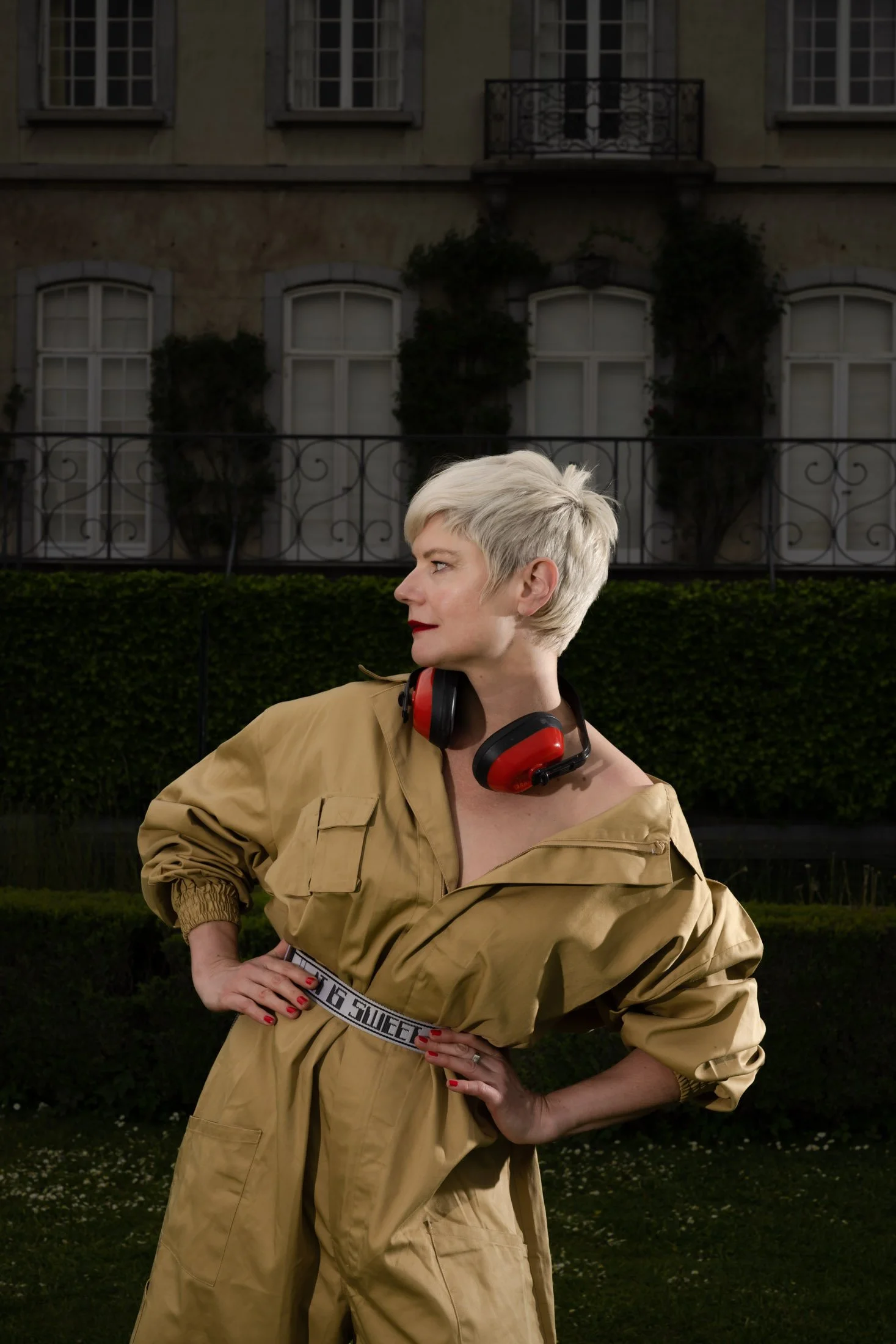 Une femme blonde avec une coiffure courte, portant un casque protecteur rouge autour du cou, se tient dans un jardin devant une maison. Elle porte une combinaison beige et a les mains sur les hanches, regardant sur le côté.