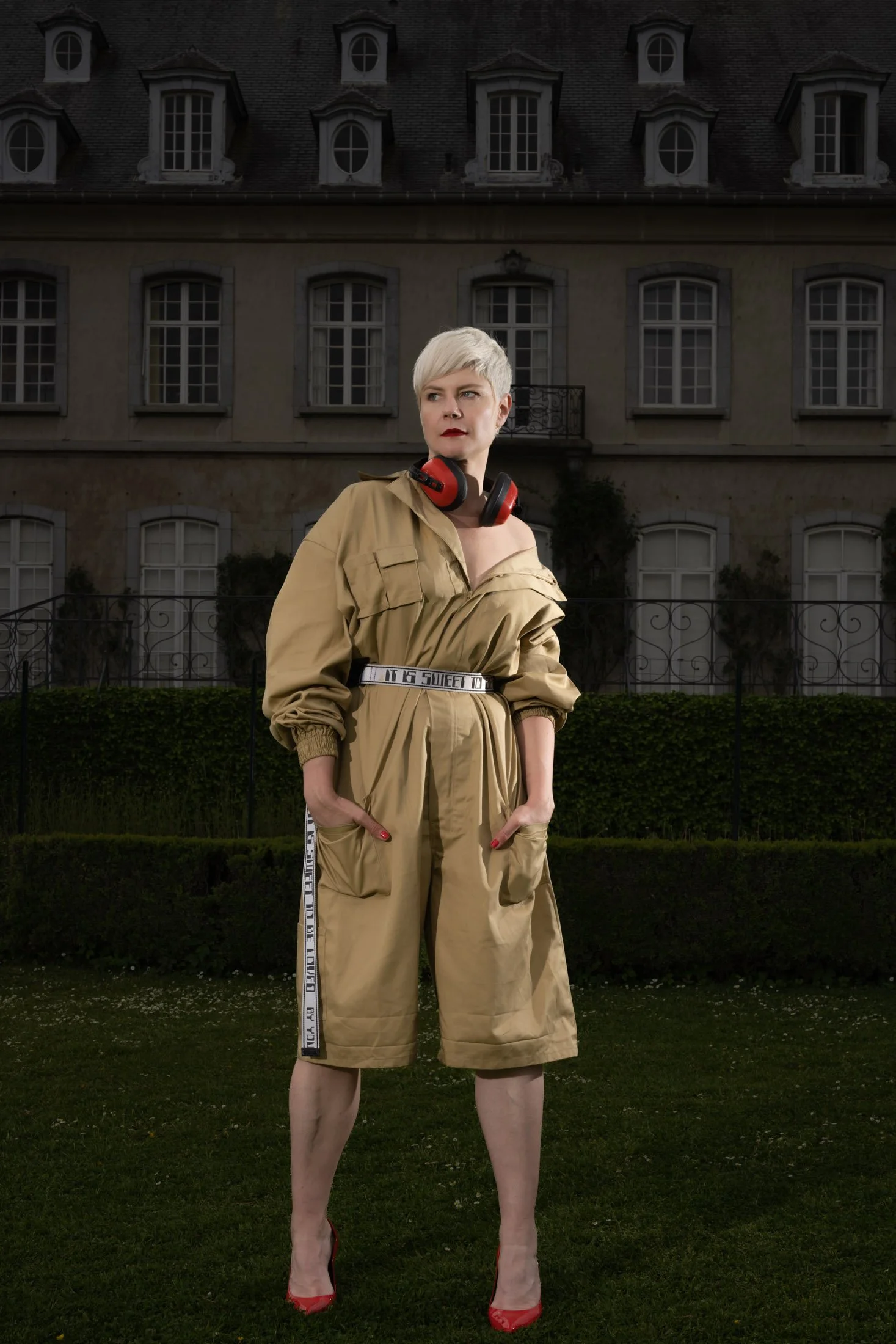 Femme aux cheveux courts en combinaison beige avec écouteurs noirs et rouges devant un bâtiment ancien.