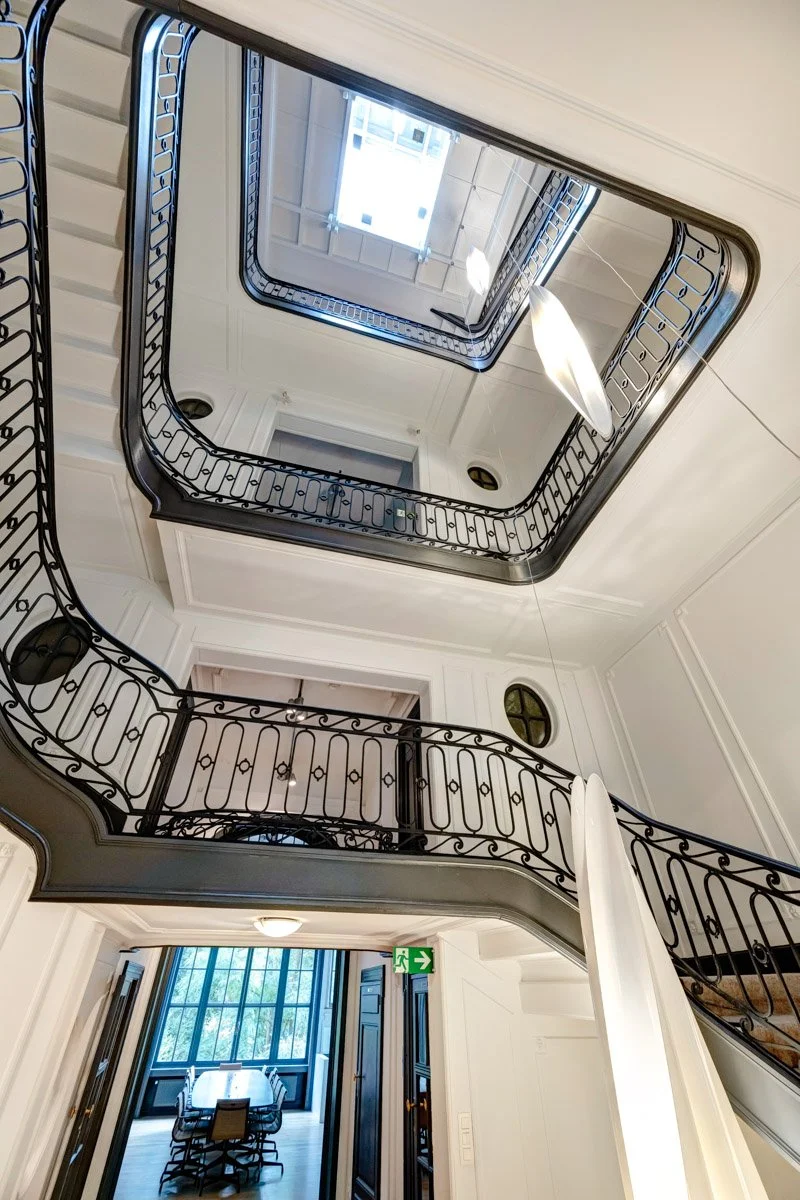 Vue intérieure d'une cage d'escalier avec des balustrades en fer forgé, montrant plusieurs étages et un plafond avec un puits de lumière.