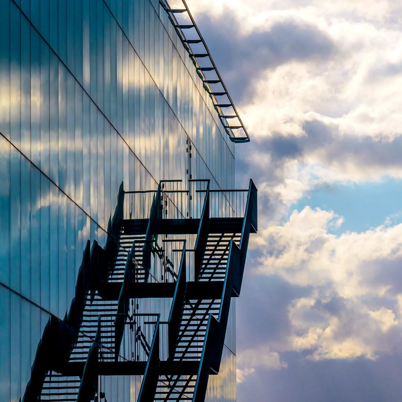Escalier de secours extérieur sur le mur d'un bâtiment moderne en verre, reflet dans la façade, avec un ciel nuageux en arrière-plan