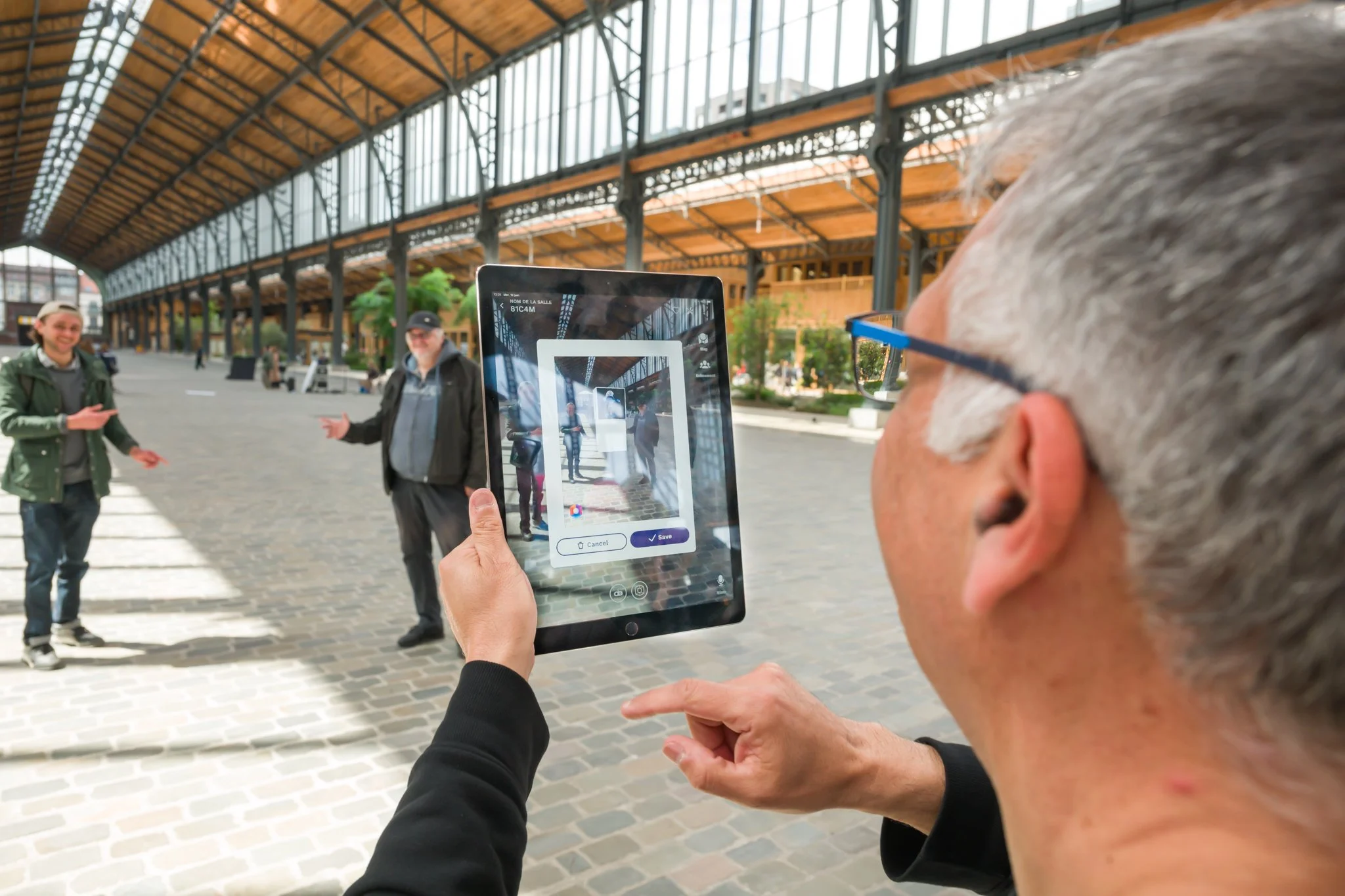 A person is using a tablet in a covered area with a paved floor and a metal and glass structure, whilst two other people are posing in the background.
