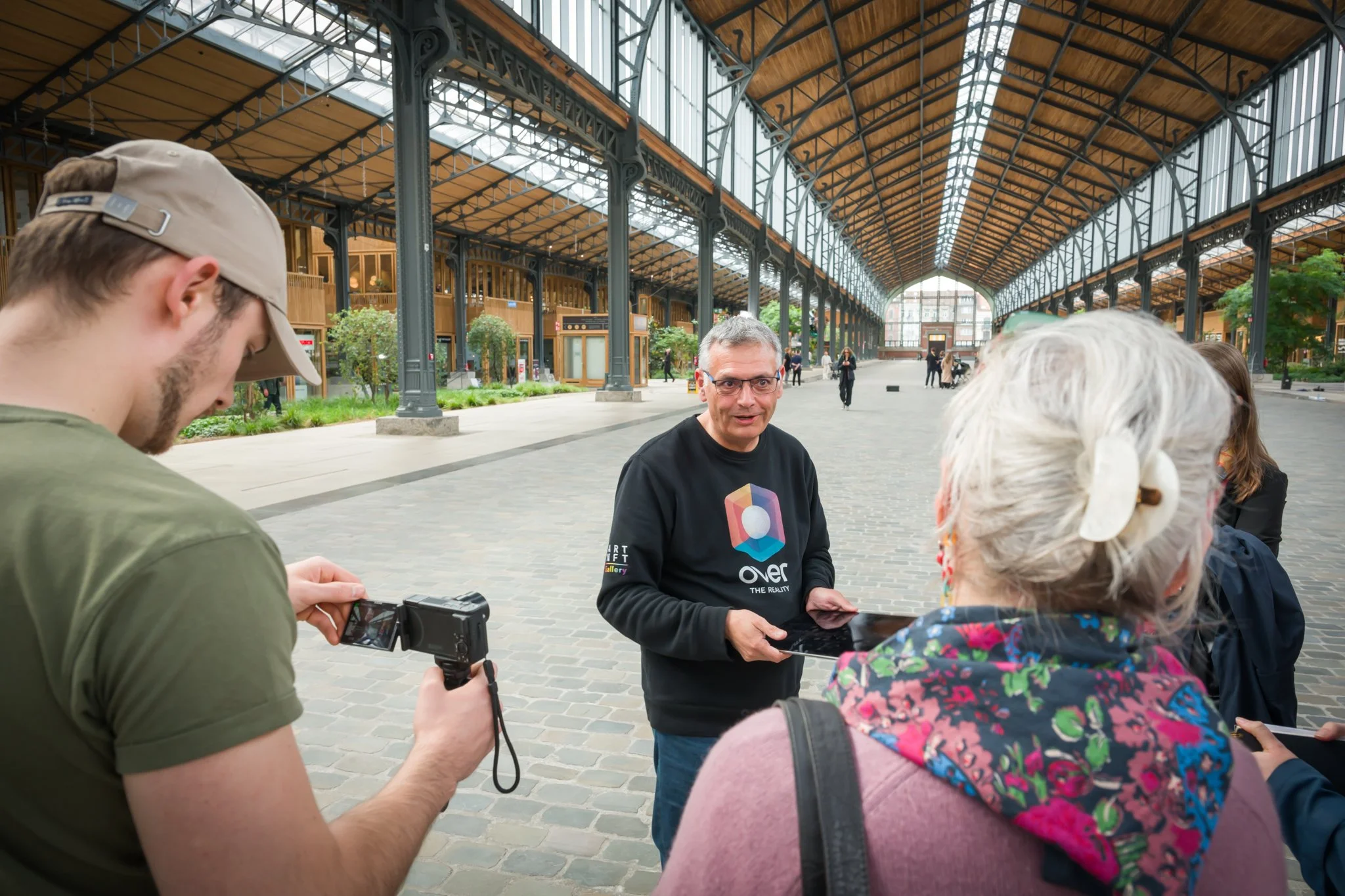 A group of people chatting in a large covered space with a steel framework and a paved floor. One person is taking a photograph of the scene, whilst another is holding a tablet. The space is bright and spacious, with a glass roof and an industrial architectural style