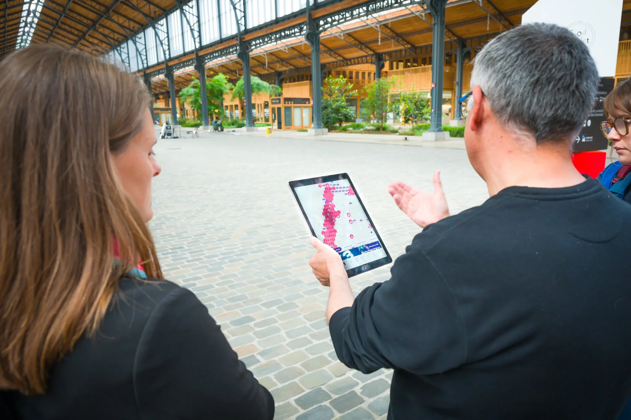 Two people looking at a tablet displaying an interactive map in a large covered hall featuring metal structures and green spaces.
