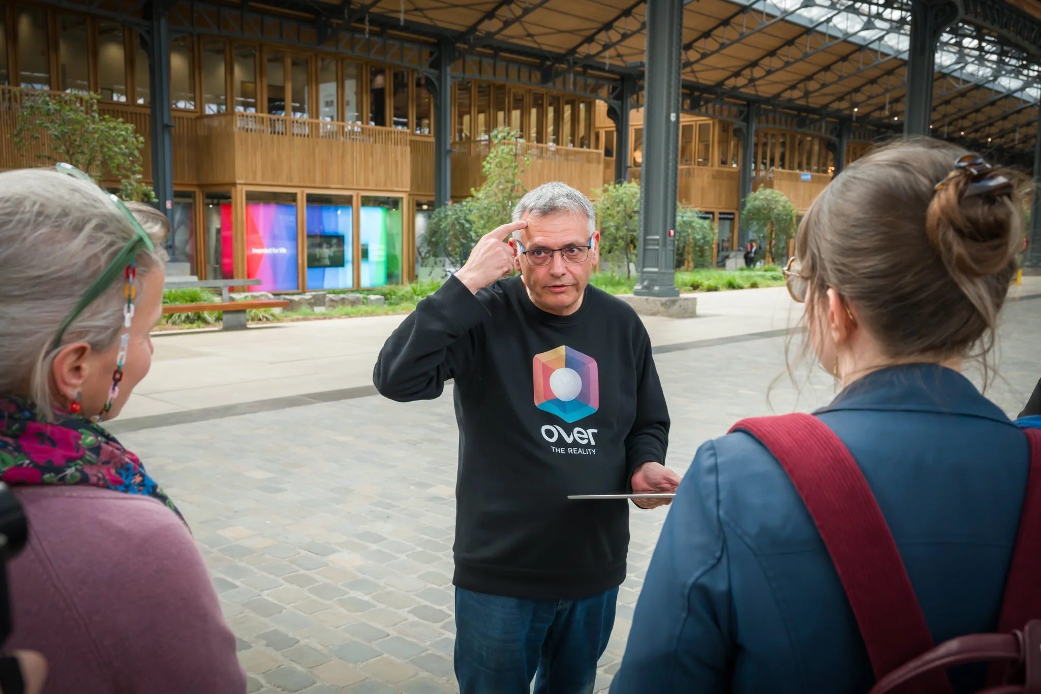 A person talking to two others in a public space, with wooden tables, plants and colourful screens in the background.