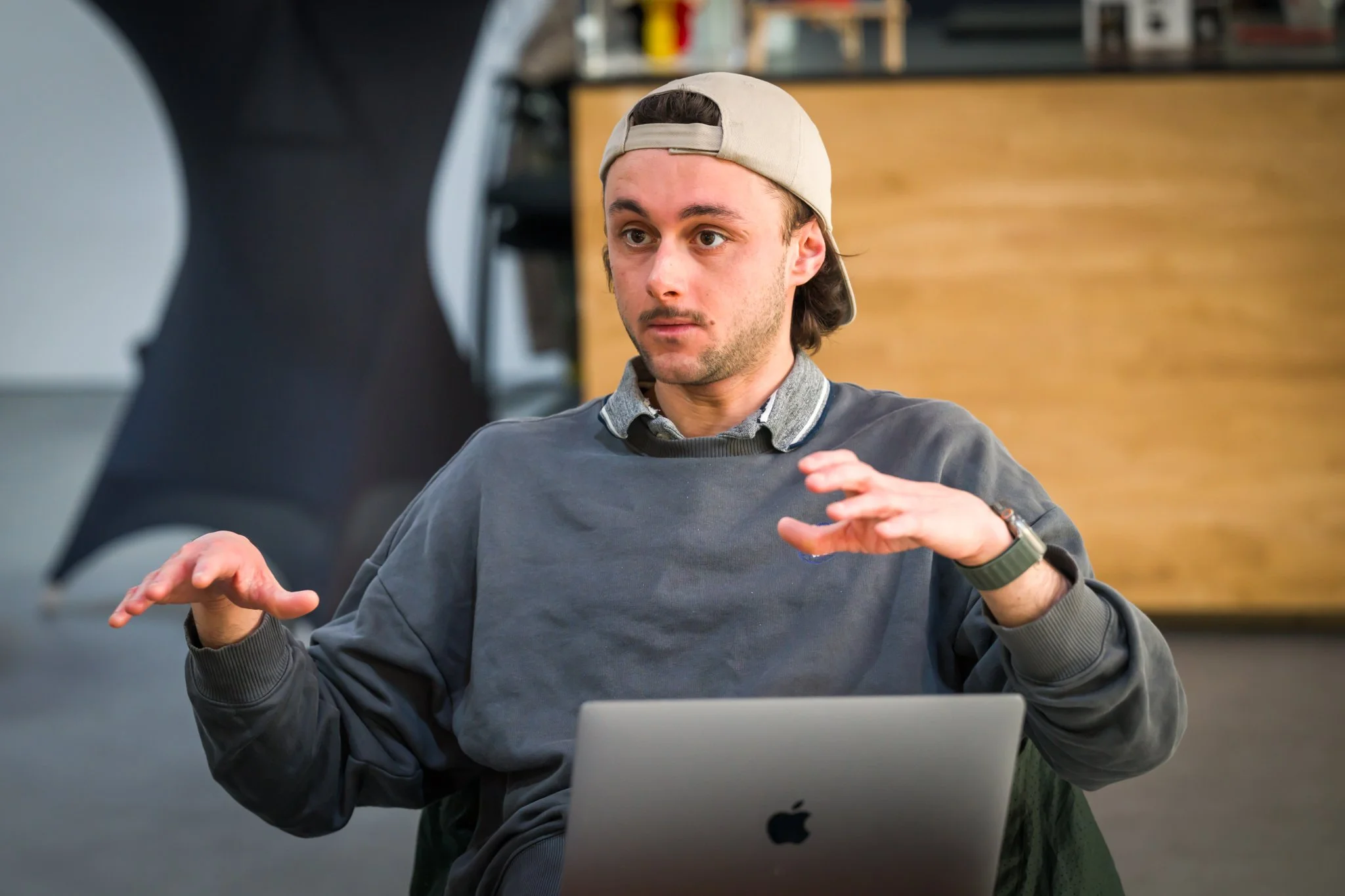 Photo d'un event à Bruxelles. Un homme portant une casquette à l'envers, vêtu d'un pull gris, est assis devant un ordinateur portable. Il semble en pleine conversation ou présentation, gesticulant avec ses mains.