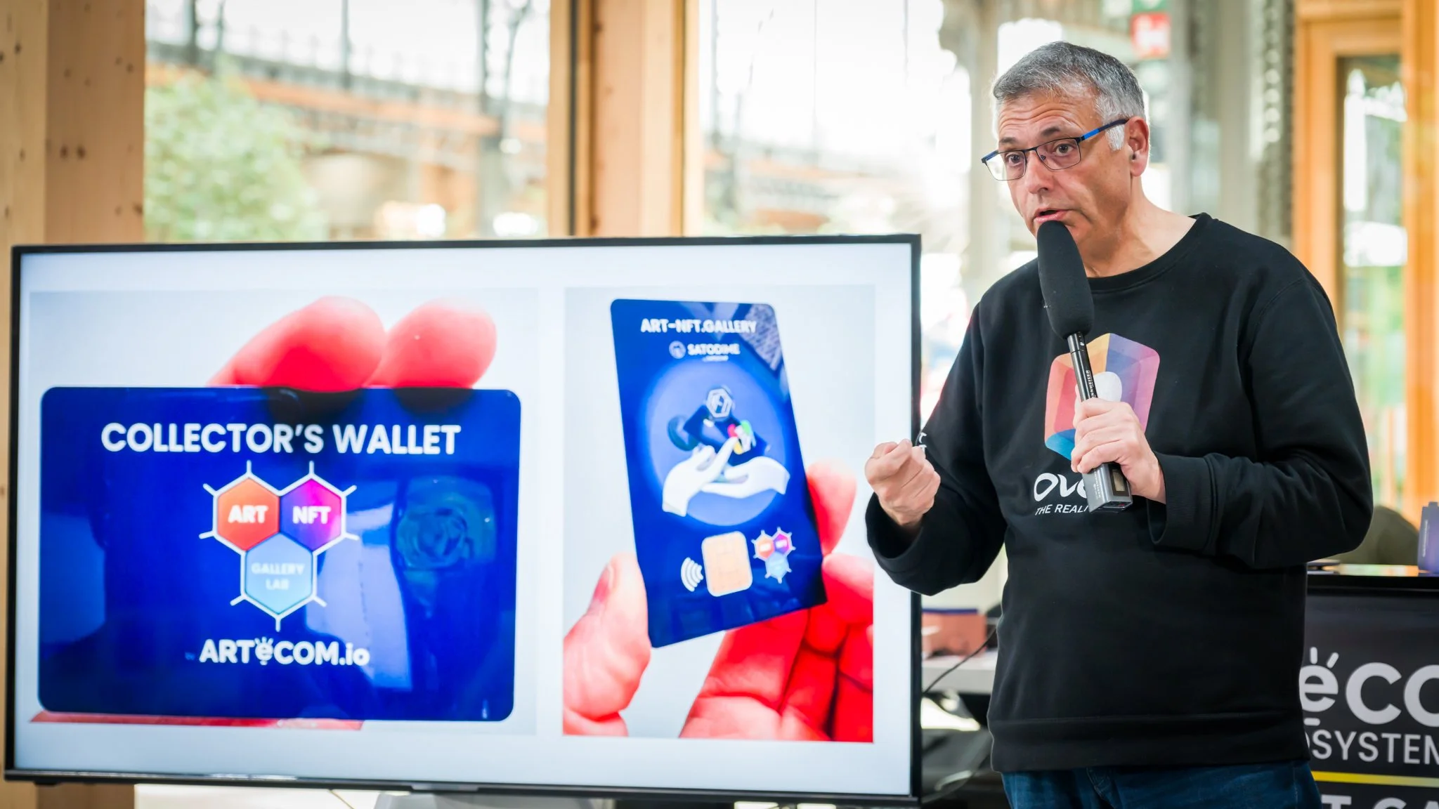 A man is holding a microphone and speaking during a presentation. There is a screen displaying digital cards entitled ‘Collector’s Wallet’, with references to NFTs and art.