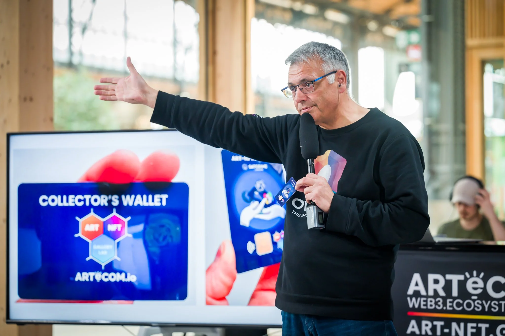 Photographie d'un événements à Bruxelles. Un homme avec un micro faisant une présentation sur les NFTs, montrant une carte devant un écran avec le texte 'Collector's Wallet'. Un public est observé à l'arrière-plan.
