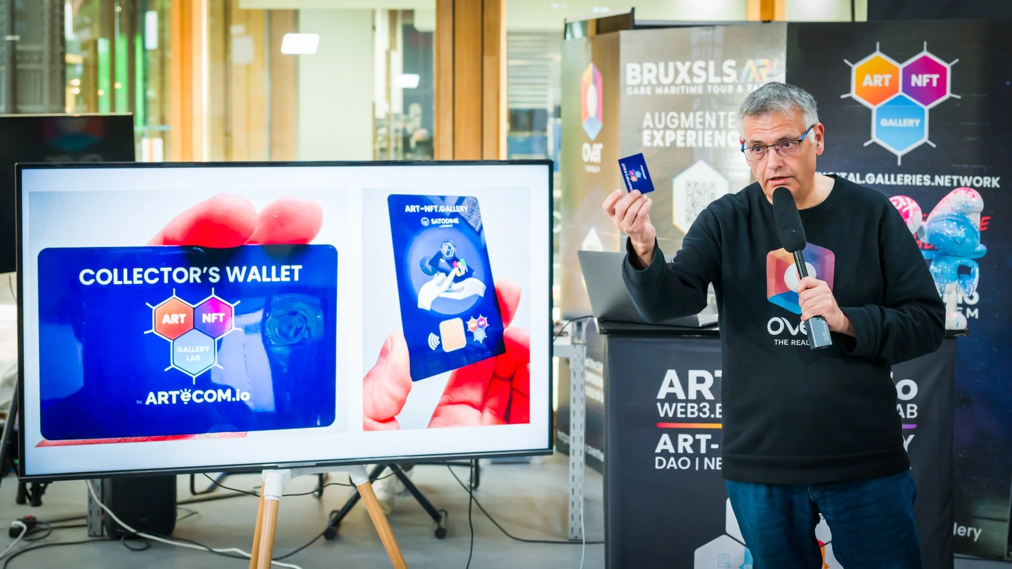 A man is holding a card and speaking into a microphone in front of a screen displaying ‘Collector’s Wallet’. There are posters about NFTs and digital art in the background.