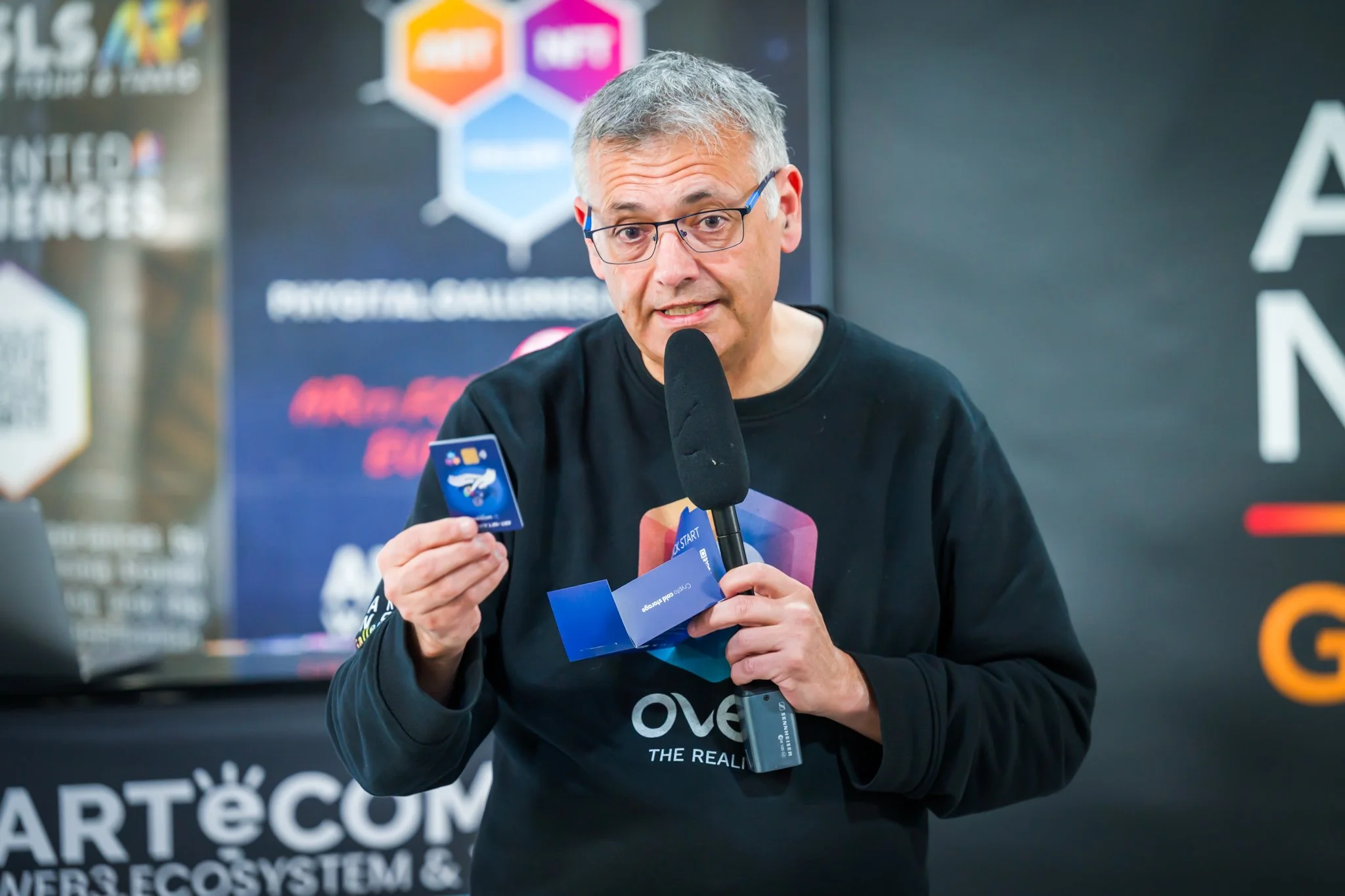 A man wearing glasses is holding a microphone and a credit card in front of a backdrop featuring logos and text about art and NFTs.