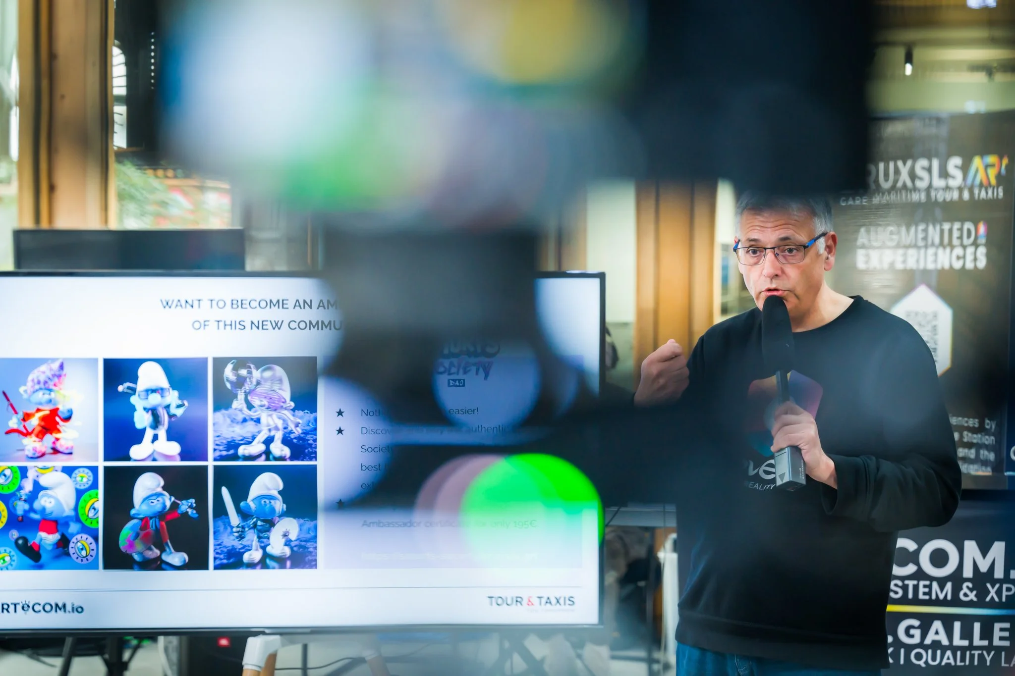 A man is speaking into a microphone in front of a screen showing images of the Smurfs. Promotional posters can be seen in the background."