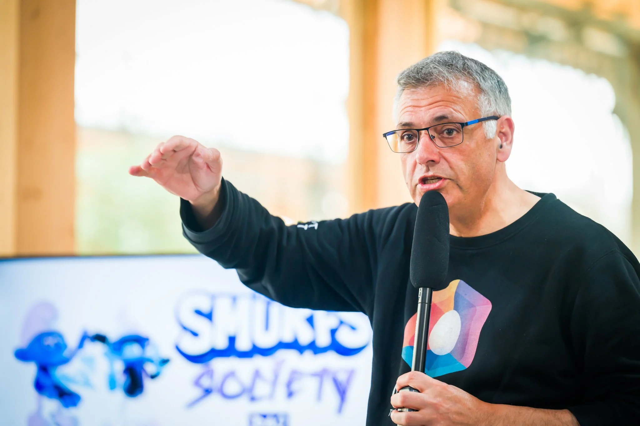 A grey-haired man wearing glasses is holding a microphone and speaking, with one hand raised. In the background, a screen displays a colourful logo. The man is wearing a jumper with a colourful geometric pattern.