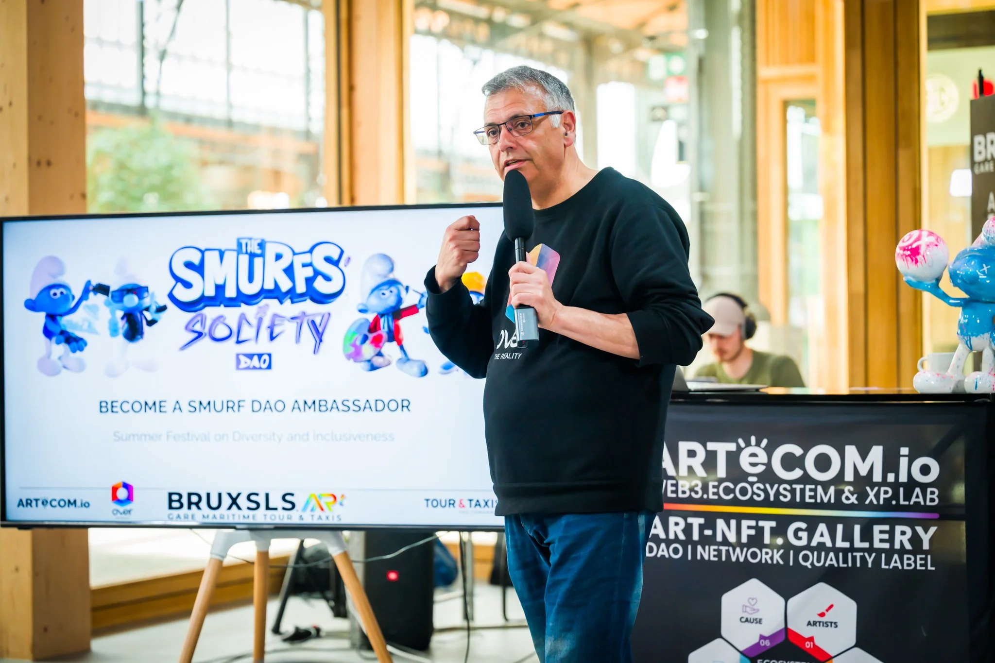 A man giving a presentation on ‘The Smurfs Society DAO’ with a microphone, in front of a screen displaying promotional material for the event and images of Smurfs. On the right, a Smurf sculpture sits on a piece of furniture alongside some adverts