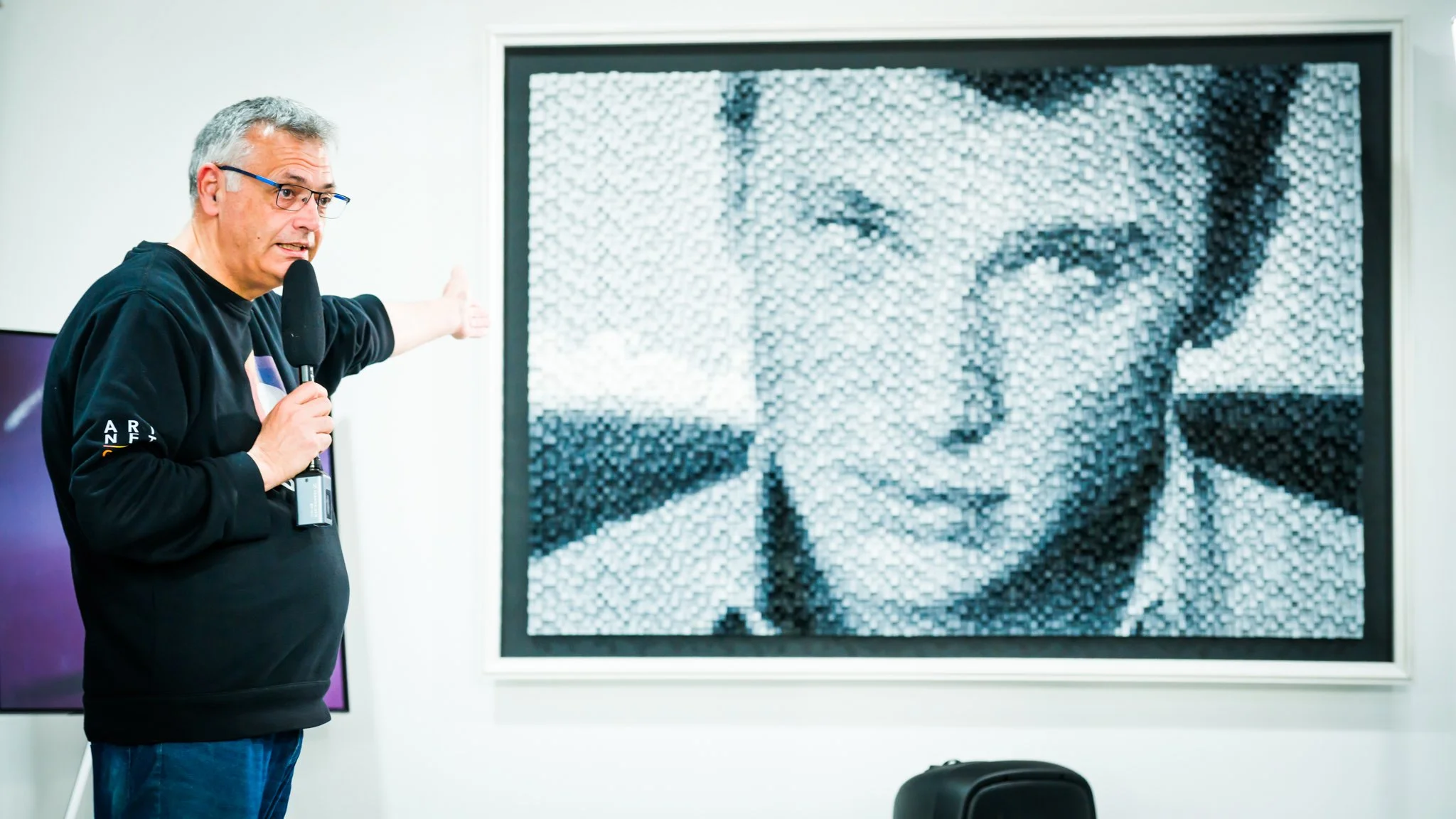 Un homme avec des lunettes, tenant un microphone, indique une peinture murale en mosaïque d'une femme souriante