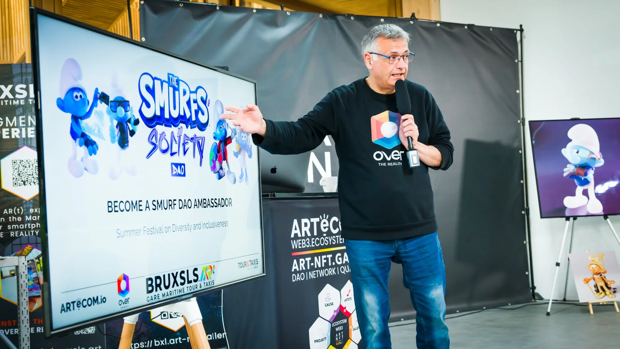 A man giving a presentation on the “Smurfs Society DAO” with a microphone, in front of a screen displaying Smurfs and the text “Become a Smurf DAO Ambassador”.
