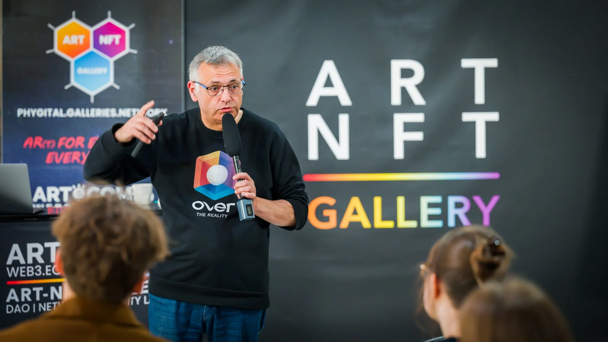 Reportage photo pendant un event d'un homme parle dans un micro lors d'un événement avec une pancarte "ART NFT GALLERY" en arrière-plan. Plusieurs personnes l'écoutent.