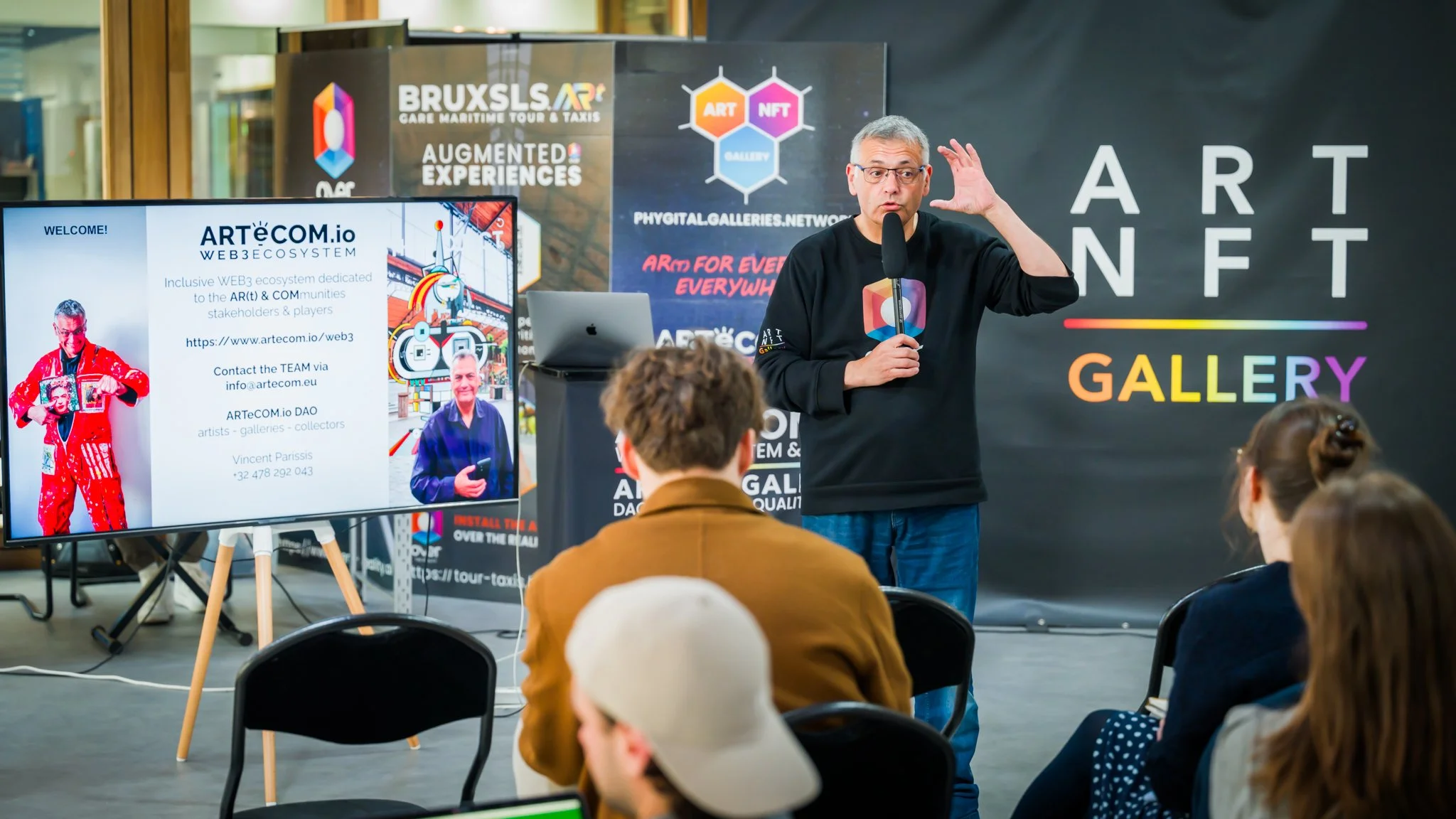 Conférencier présentant devant un public avec écran affichant 'ARTéCOM.io', décor NFT et galerie d'art.