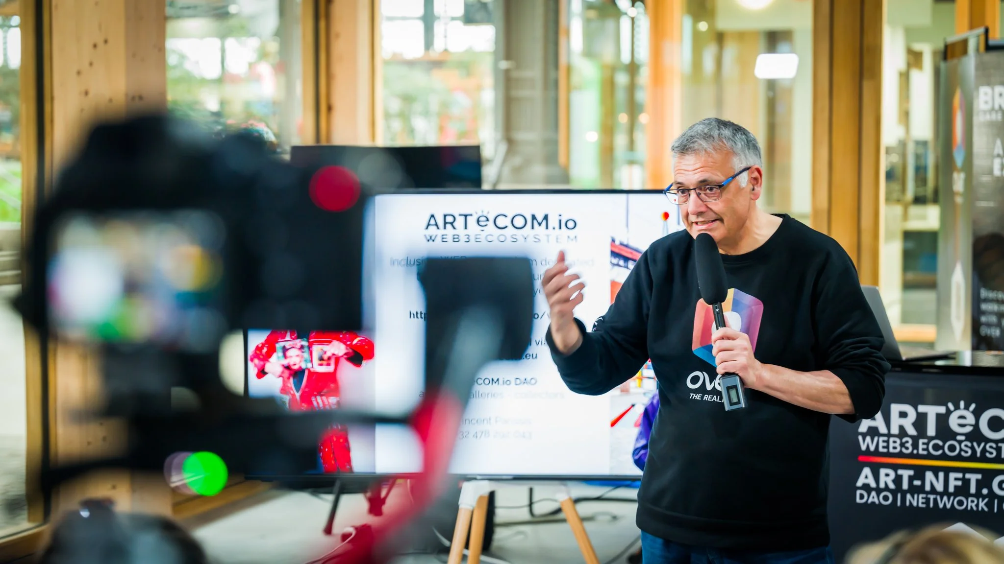 Un homme parle lors d'une présentation en intérieur avec un micro, devant un écran et une bannière affichant 'ARTéCOM.io'.