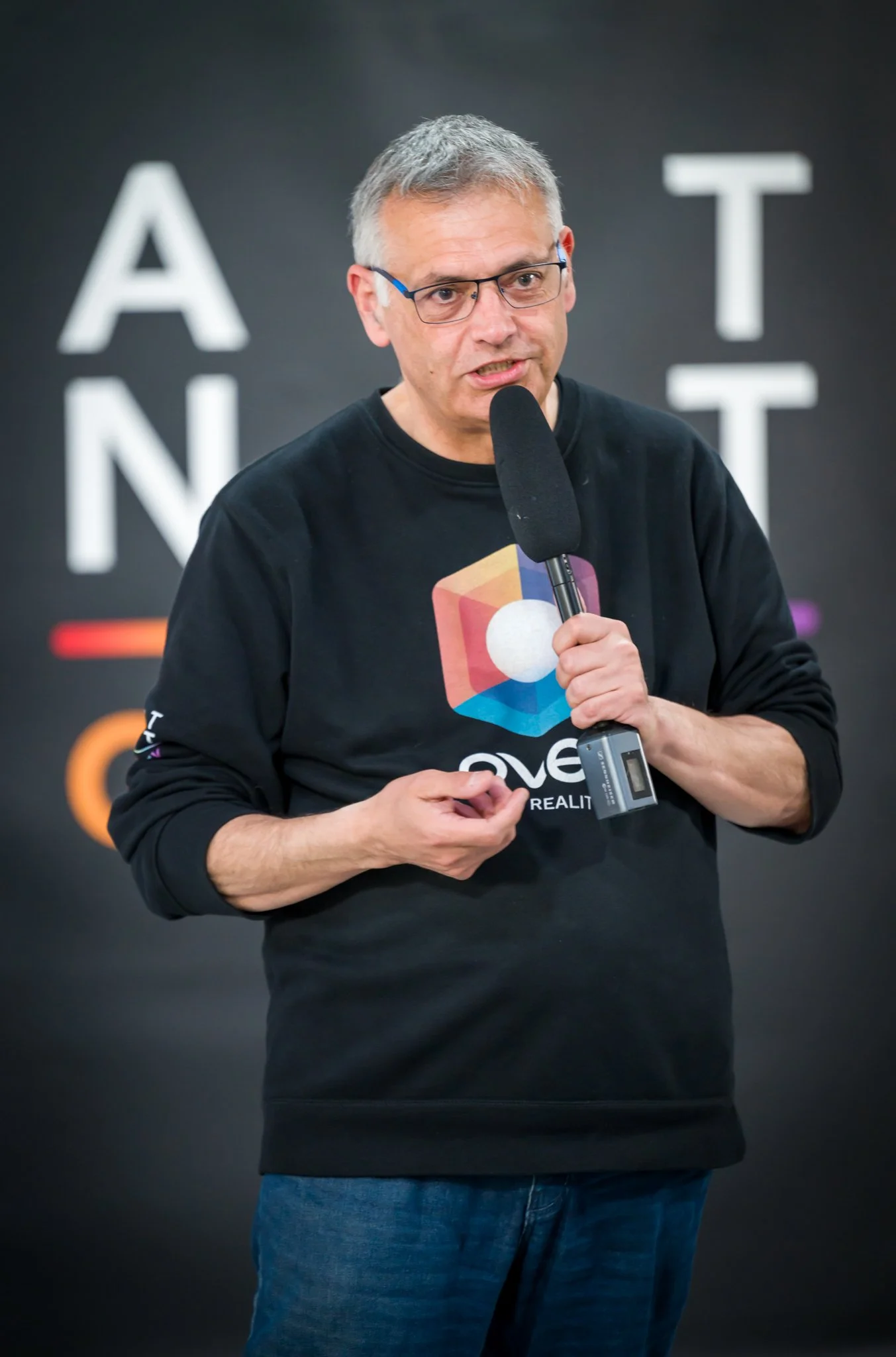 A man holding a microphone, wearing a black jumper with a logo, in front of a blurred background with white letters.