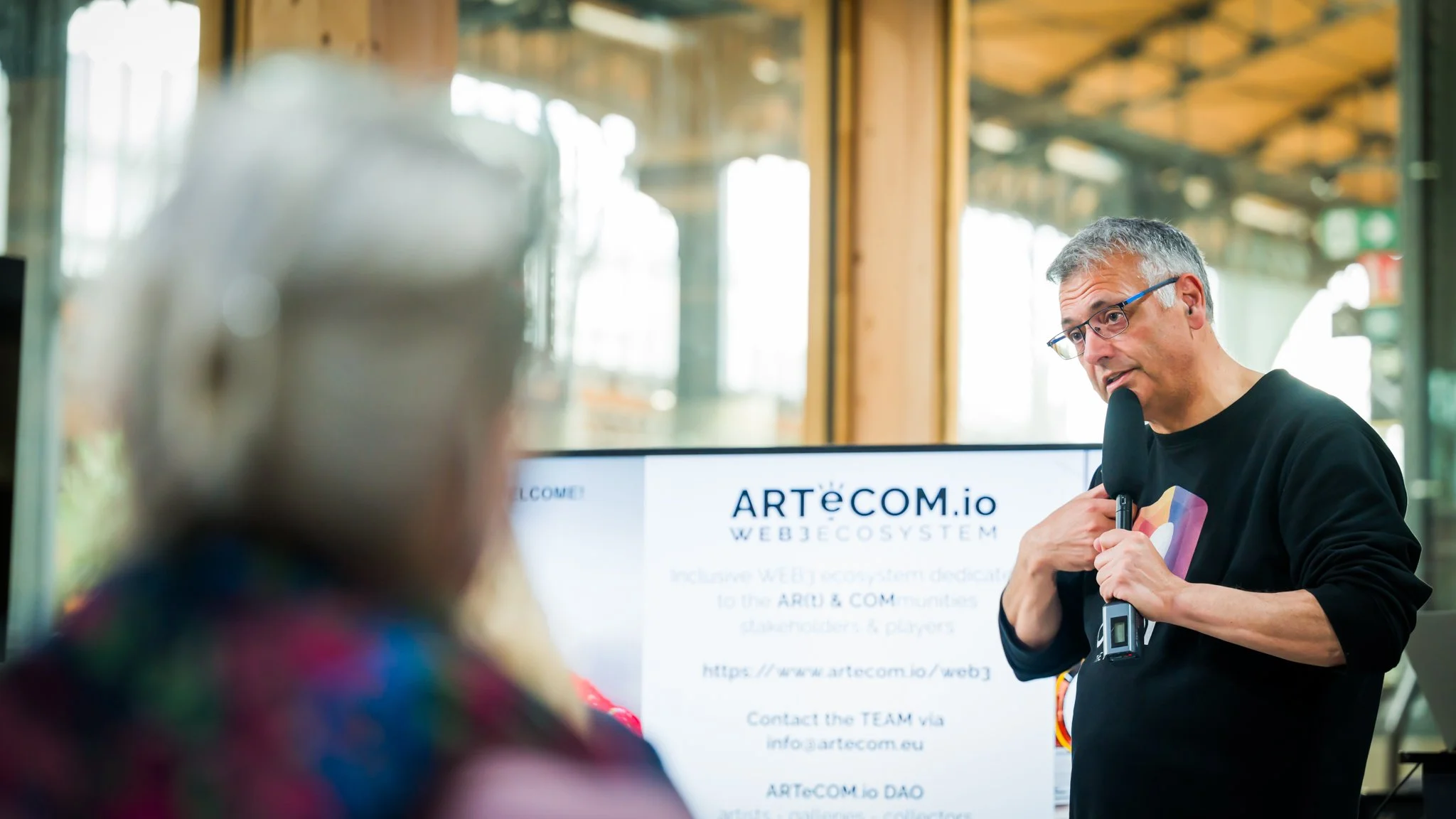 A man speaking into a microphone in front of a screen displaying the website ‘ARTeCOM.io’. A person, seen from behind, is listening. A well-lit setting with wooden structures visible in the background.