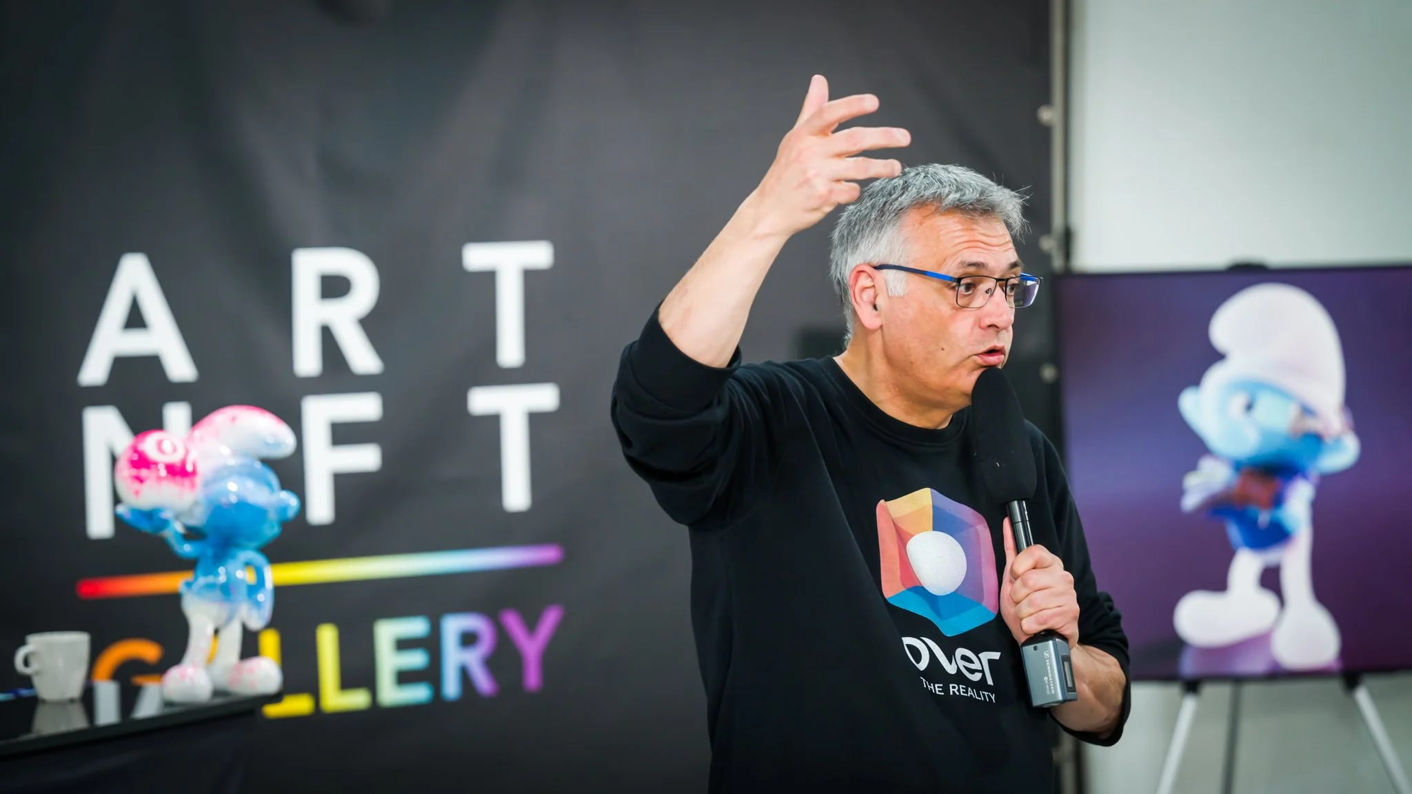 A man wearing glasses is speaking into a microphone at an event. There is a poster reading ‘ART NFT GALLERY’ and a Smurf sculpture or figurine on a table. In the background, an image of a Smurf is projected onto a screen.