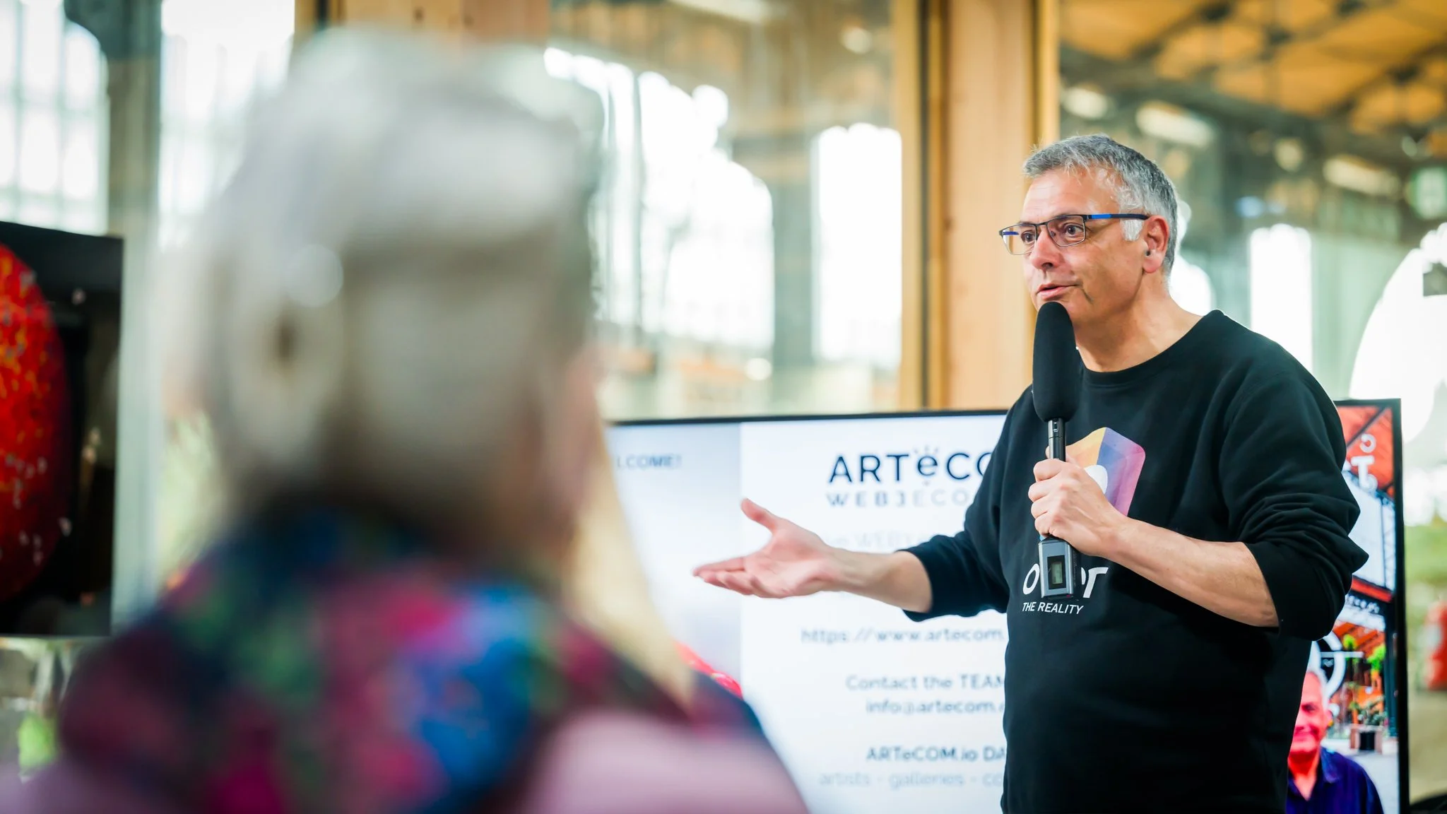 A man speaking into a microphone in front of a screen displaying ‘ARTeCOM’, with a person seen from behind in the foreground. The setting appears to be an indoor location with large windows in the background.