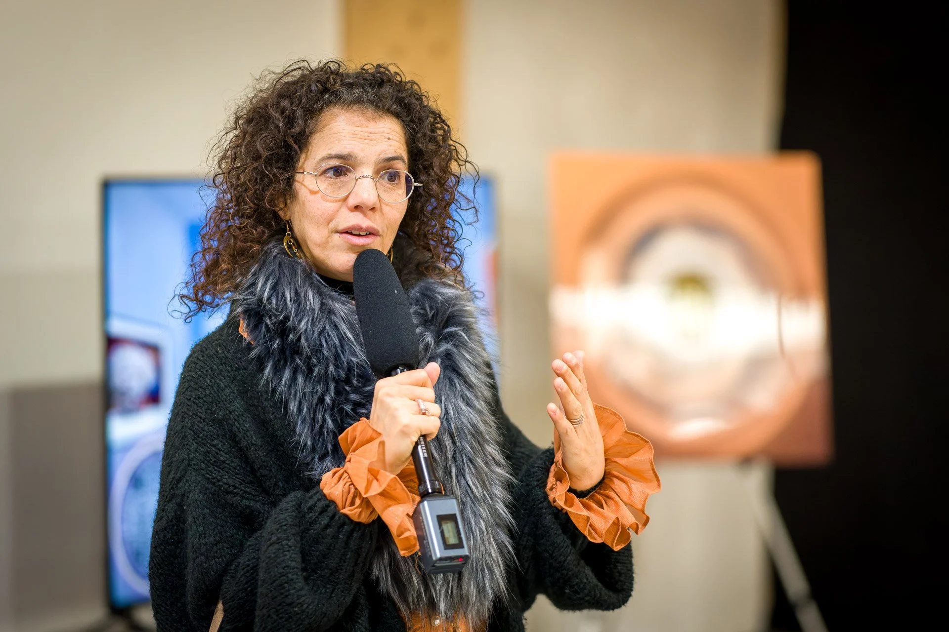 Hanane Risayindi s’exprime au microphone, avec des écrans et une œuvre à l’arrière-plan, à la BXL ART NFT Gallery.