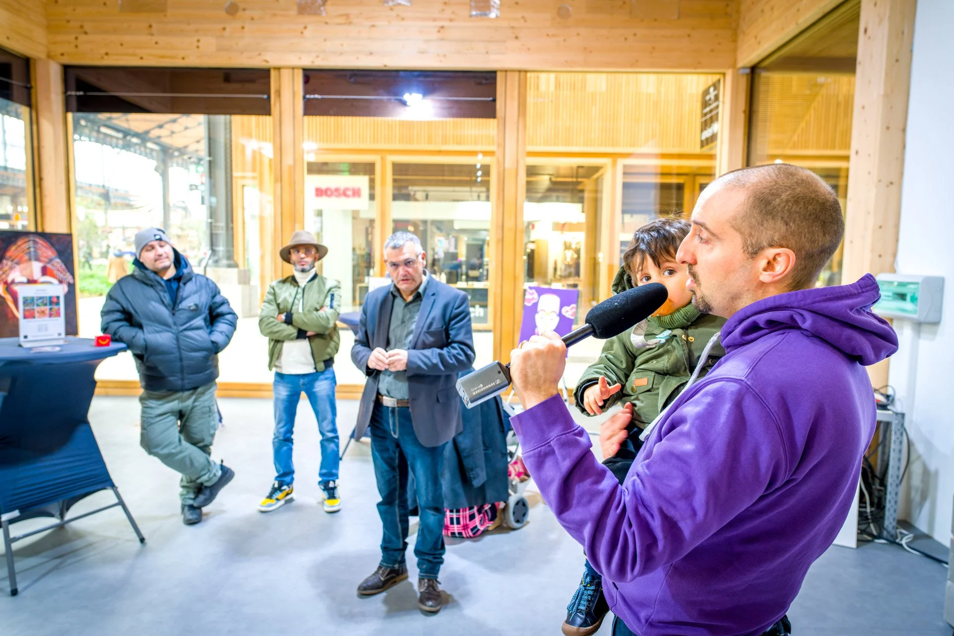 Un homme en sweat à capuche violet tenant un enfant et parlant dans un microphone, devant un petit groupe de personnes dans un espace intérieur avec des fenêtres en bois et une affiche. Les gens regardent attentivement.