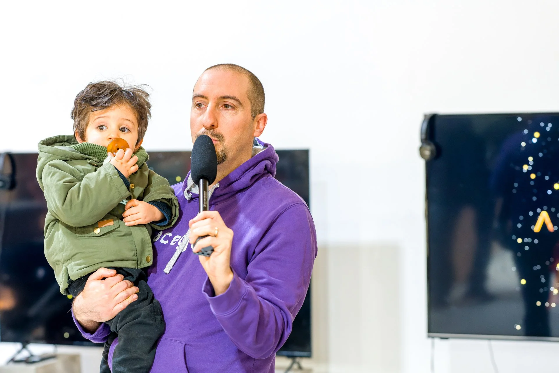 Un homme en pull violet tenant un enfant portant une veste verte. L'homme parle dans un microphone. En arrière-plan, un écran visiblement allumé.