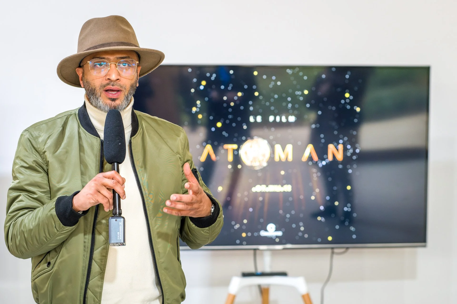Un homme avec un manteau vert et un chapeau parle dans un micro devant un écran affichant le titre 'Atoman'. L'écran montre un arrière-plan étoilé.