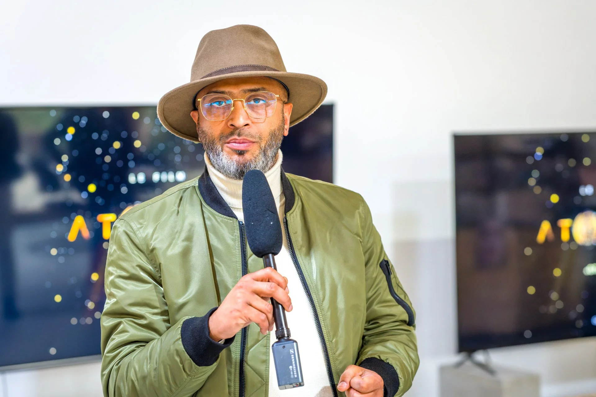 Un homme portant un chapeau et des lunettes tient un microphone devant des écrans.