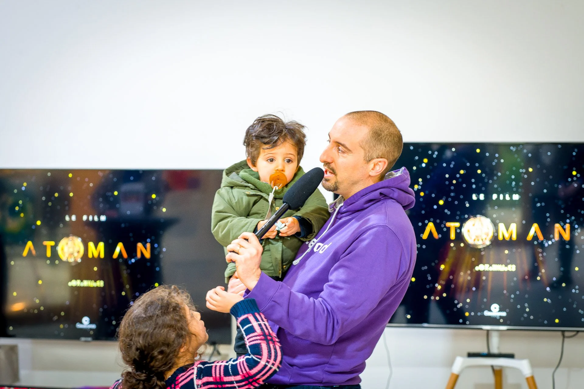 Un homme en sweat à capuche violet tient un enfant avec une sucette et un microphone, devant deux écrans affichant "ATOMAN".